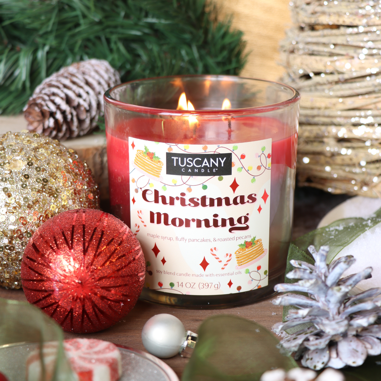 A lit Tuscany Candle® SEASONAL "Christmas Morning Scented Holiday Candle" (14 oz) sits among ornaments, pinecones, and greenery for a festive holiday display.