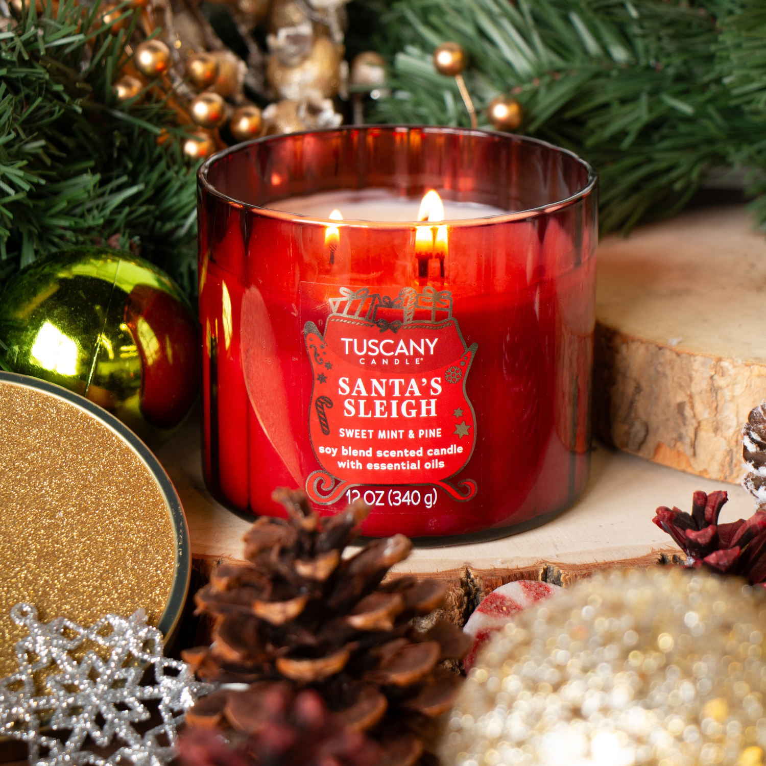 A lit 12 oz Tuscany Candle® SEASONAL "Santa's Sleigh: Sweet Mint & Pine" from the Winter Icons Collection rests on wood, surrounded by pine cones, ornaments, and evergreens.