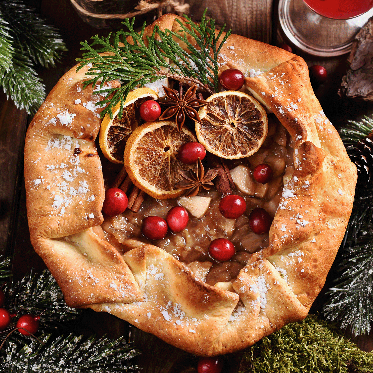 A rustic pie garnished with dried orange, cranberries, and star anise rests among pine sprigs, capturing the festive aroma of Tuscany Candle® SEASONAL Holiday Sips: Winter Sangria Scented Holiday Candle (12 oz) from the Winter Icons Collection.