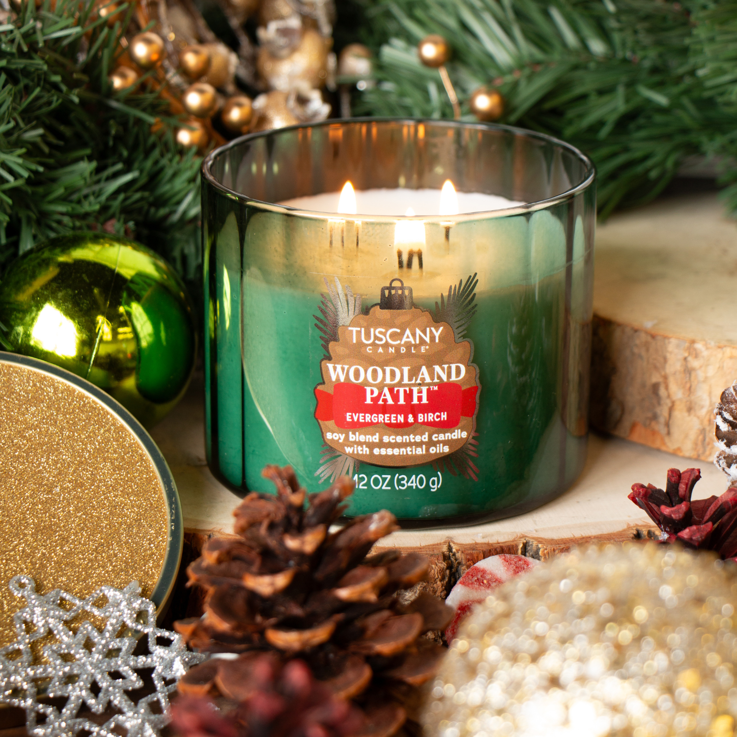 A lit Tuscany Candle® SEASONAL "Woodland Path: Evergreen & Birch Scented Holiday Candle" (12 oz) from the Winter Icons Collection glows among pine cones, ornaments, and evergreen branches in a festive holiday scene.