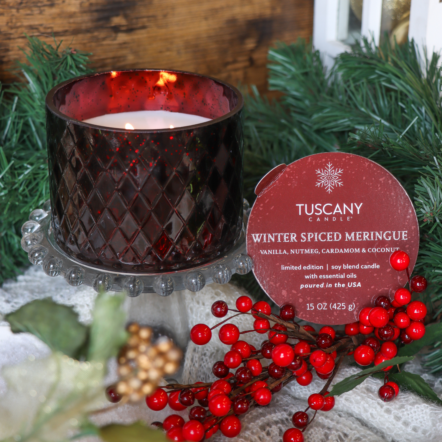 A 15 oz Tuscany Candle® SEASONAL "Winter Spiced Meringue: Spicy Cardamom & Vanilla Cream" holiday candle from the Winter Glow Collection sits on a glass stand, surrounded by faux greenery and red berries.
