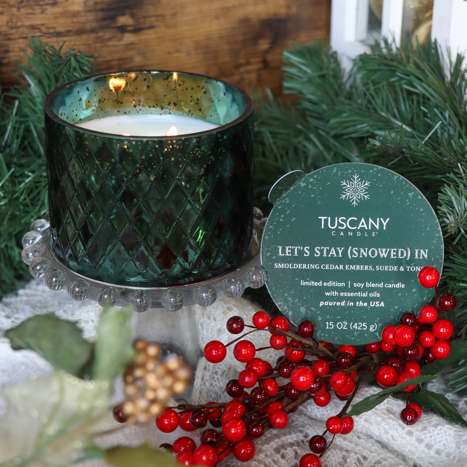 A 15 oz Tuscany Candle® SEASONAL “Let’s Stay (Snowed) In: Charred Firewood & Black Pepper” candle from the Winter Glow Collection sits on a glass stand beside red berries, greenery, and winter decor.