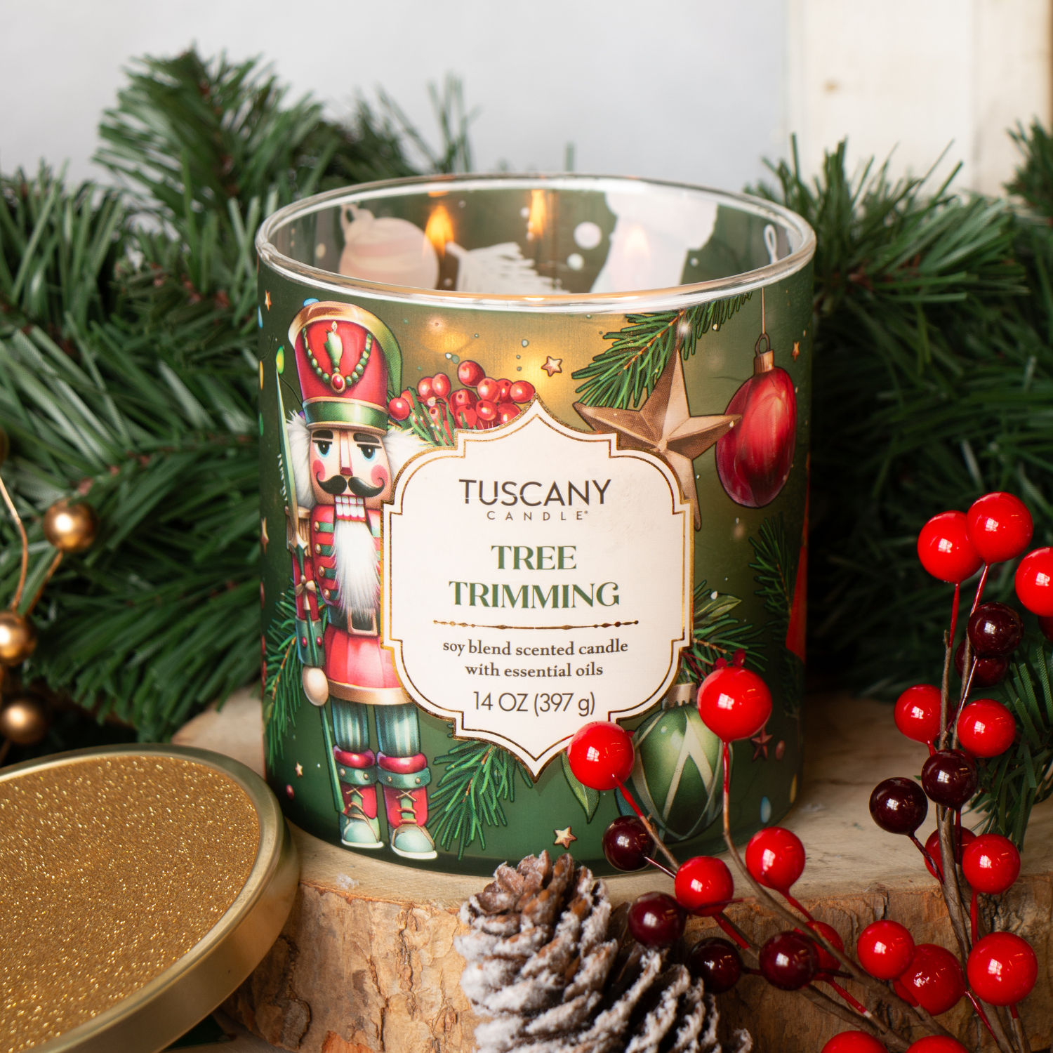 A 14 oz Tuscany Candle® SEASONAL "Tree Trimming: Frosted Fir & Juniper" candle from the Traditions Collection sits in a festive glass jar on wood, surrounded by pine, berries, and a pine cone. The lid is set aside.