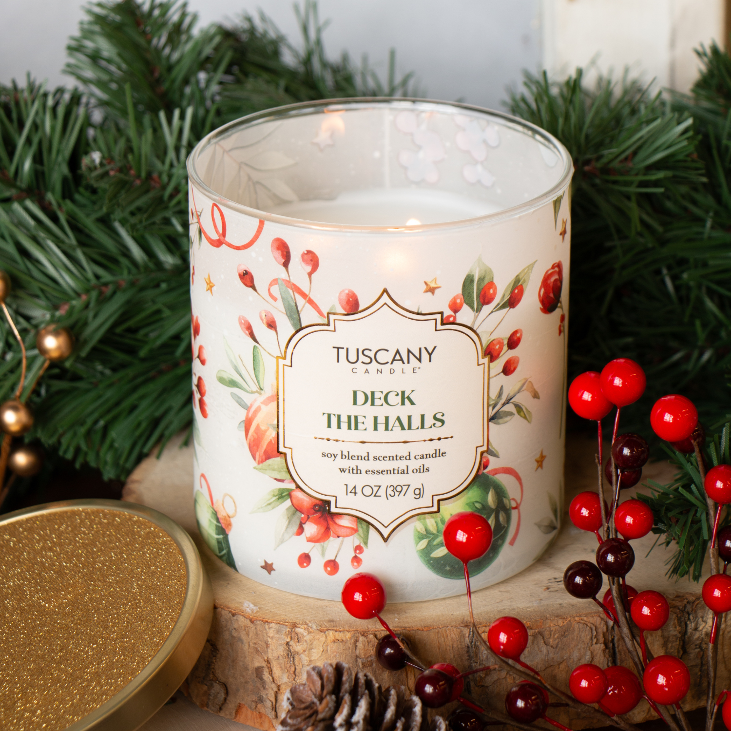A Tuscany Candle® SEASONAL "Deck The Halls: Evergreen & Clementine" 14 oz candle from the Traditions Collection sits on a table with festive greenery, red berries, and its gold lid nearby.