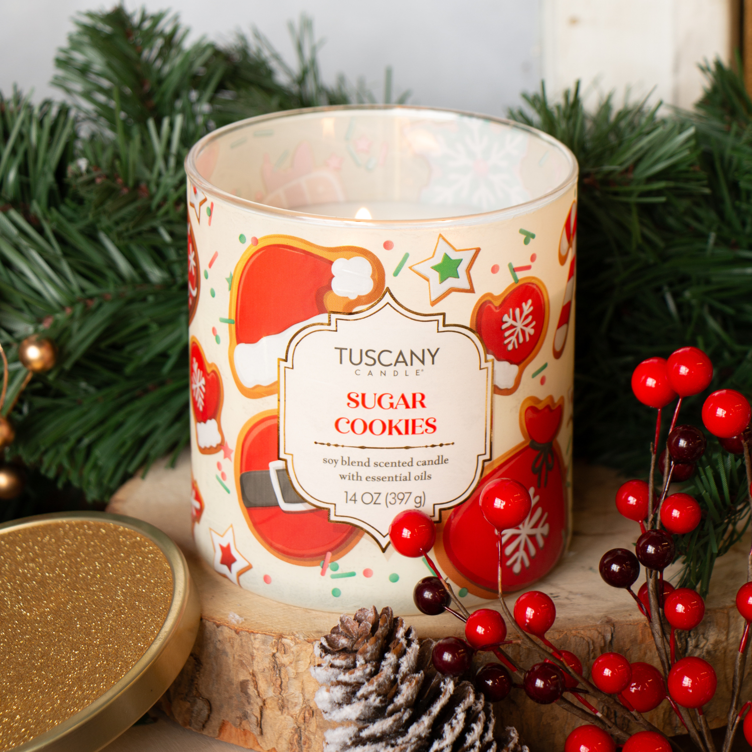A lit Tuscany Candle® SEASONAL Sugar Cookies: Buttercream Frosted Cookie Scented Holiday Candle (14 oz) from the Traditions Collection sits among pine branches, pine cones, red berries, and Christmas decor, with its festive lid beside it.