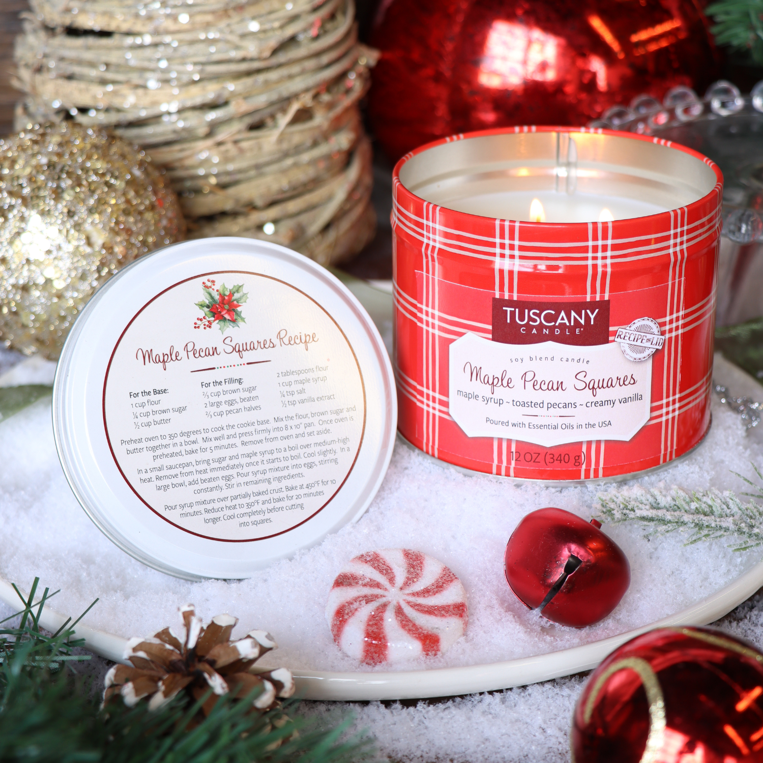 A Tuscany Candle® SEASONAL 12 oz tin—Maple Pecan Squares: Maple & Cinnamon Pancake Scent, part of the Bakery Tins Collection—sits open on snow, showing its recipe, with Christmas ornaments and greenery nearby.