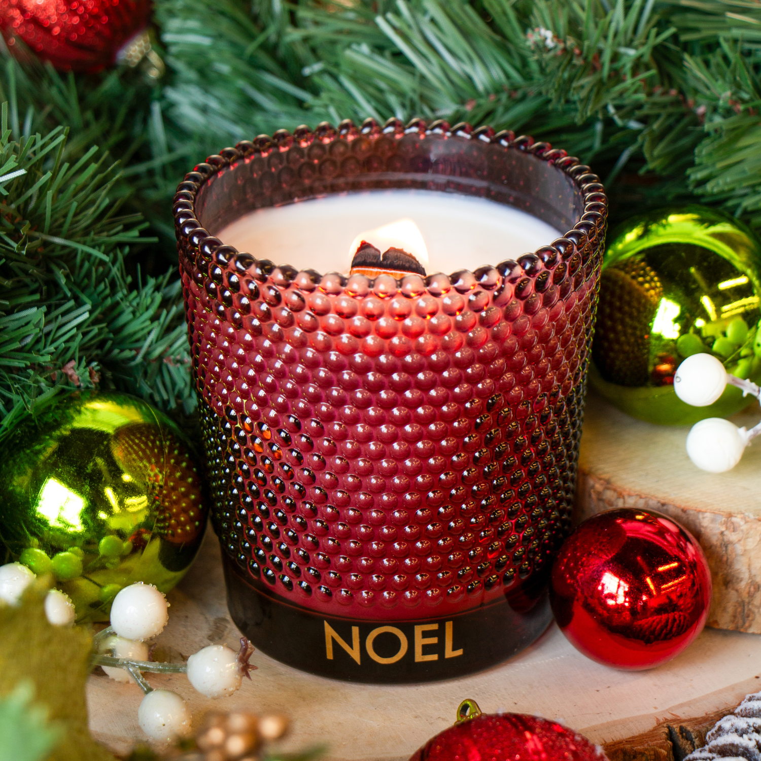 The Tuscany Candle® SEASONAL Noel: Festive Sangria Scented Holiday Wooden Wick Candle (13 oz) glows among pine branches and Christmas ornaments on a wooden surface.