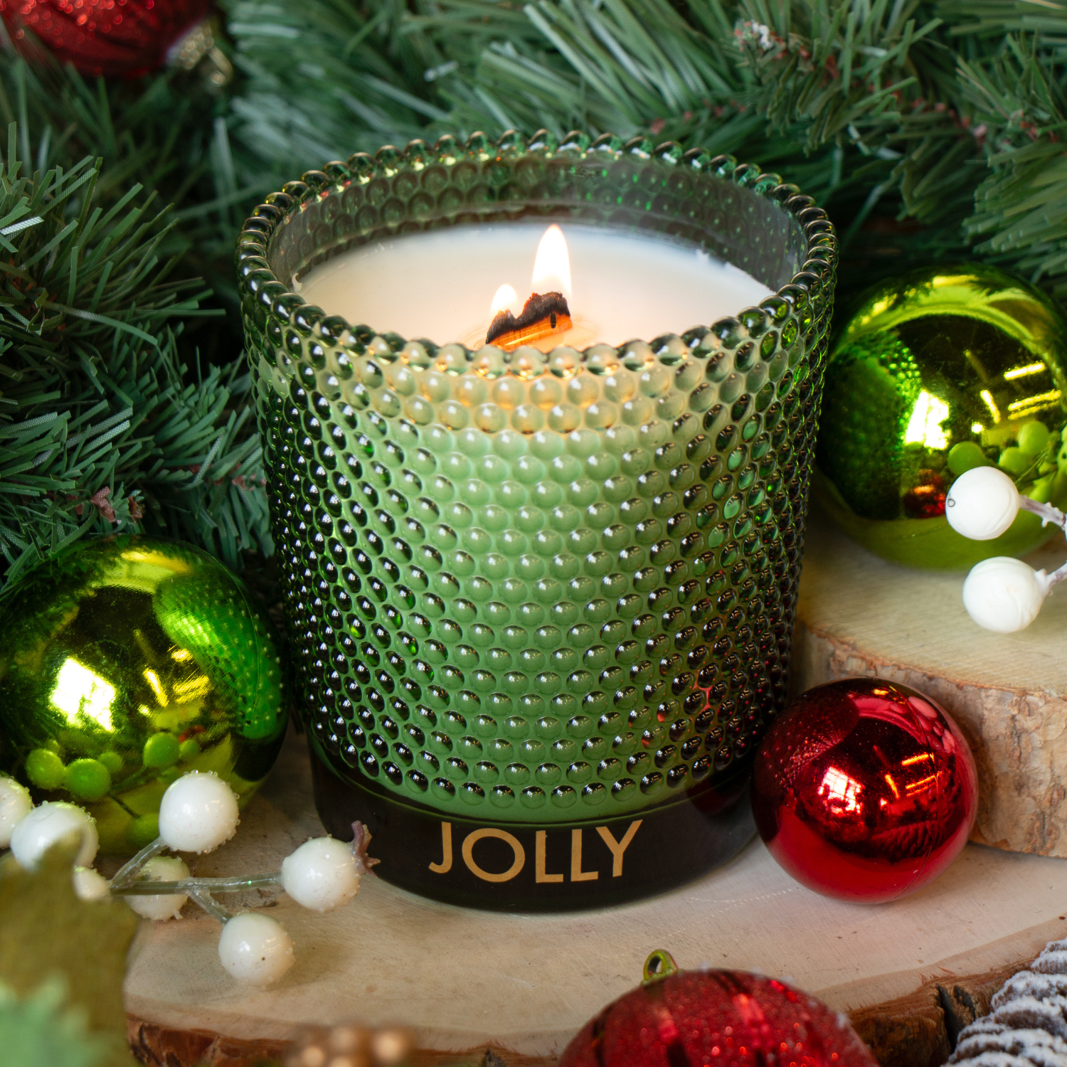 A Tuscany Candle® SEASONAL Jolly: Spicy Clove & Frosted Spruce 13 oz wooden wick candle glows in a textured glass holder, surrounded by Christmas ornaments, pine branches, and white berries.