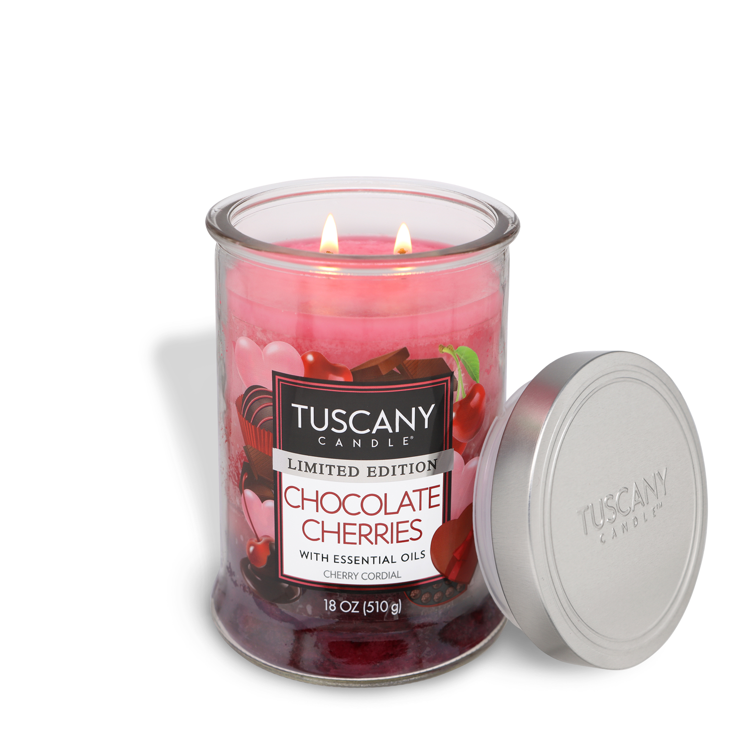 A lit Tuscany Candle® SEASONAL 18 oz glass jar labeled "Chocolate Cherries: Cherry Cordial Scented Valentine's Candle" sits with its silver lid beside it. The pink candle features cherry and chocolate images, perfect for Valentine’s Day.