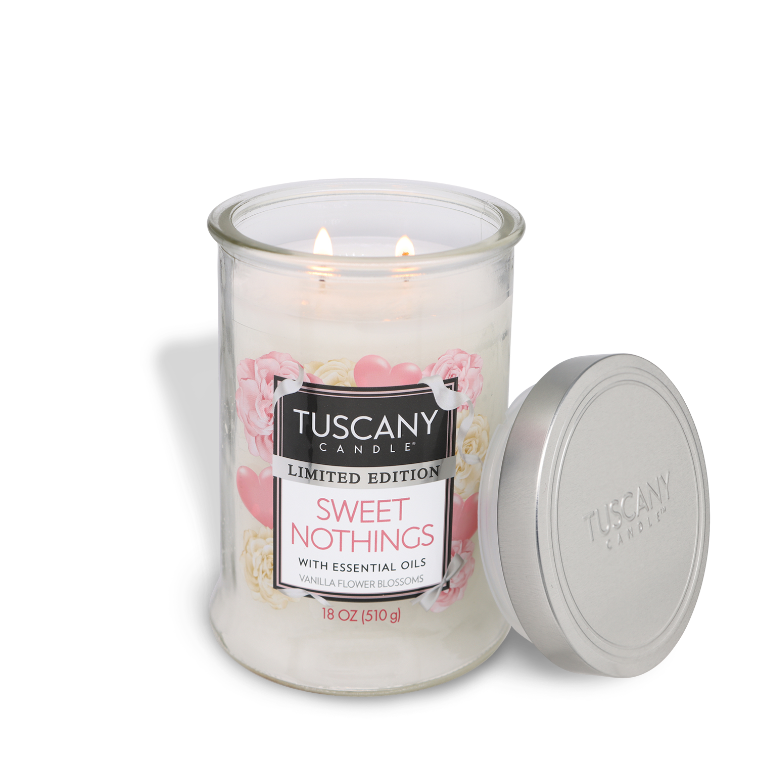 A Tuscany Candle® SEASONAL 18 oz "Sweet Nothings: Violet & Magnolia" candle with essential oils burns with two flames, its silver lid placed beside the glass jar.