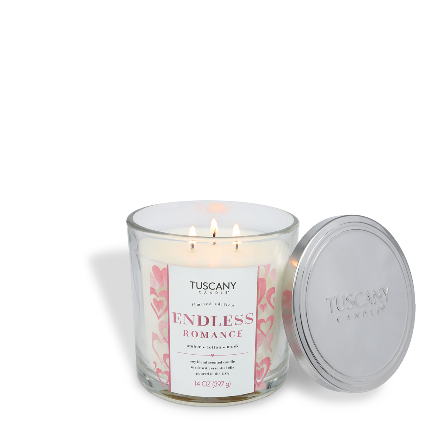 A Tuscany Candle® SEASONAL "Endless Romance: Amber & Musk Scented Valentine's Candle" (14 oz) featuring pink heart designs burns beside its silver lid on a white surface.