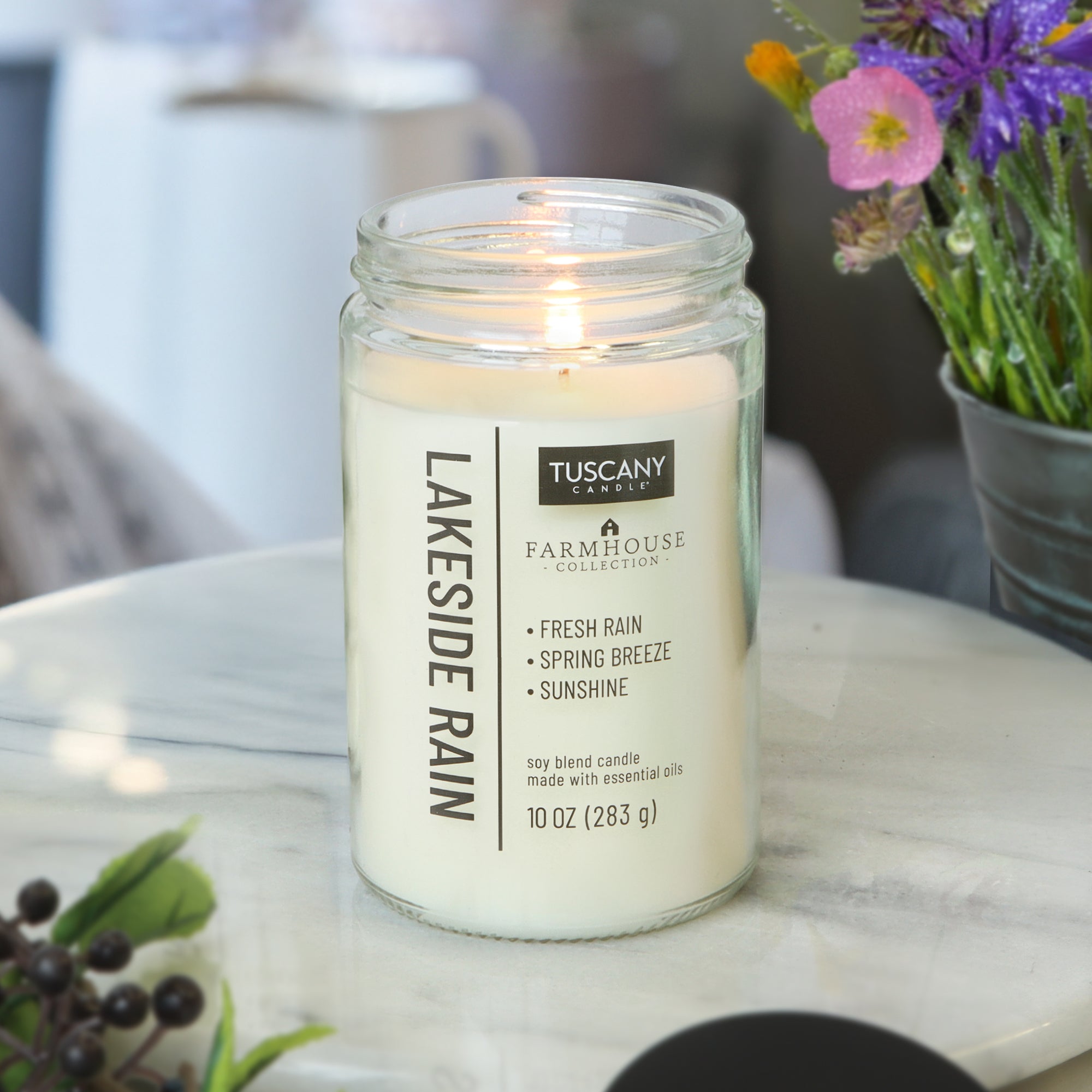 A lit Tuscany Candle® EVD Lakeside Rain Scented Jar Candle (10 oz) from the Farmhouse Collection sits on a marble table with flowers and berries, making it an ideal accent for farmhouse decor.