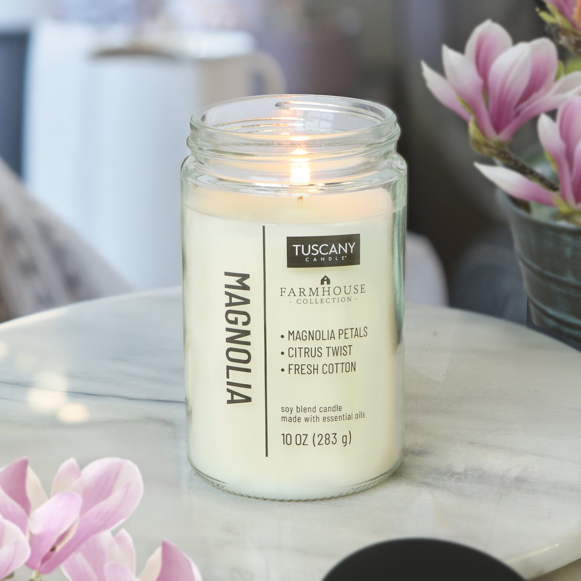 A lit Magnolia Scented Jar Candle (10 oz) from the Tuscany Candle® EVD Farmhouse Collection sits on a marble surface beside pink magnolia flowers in a softly lit room.