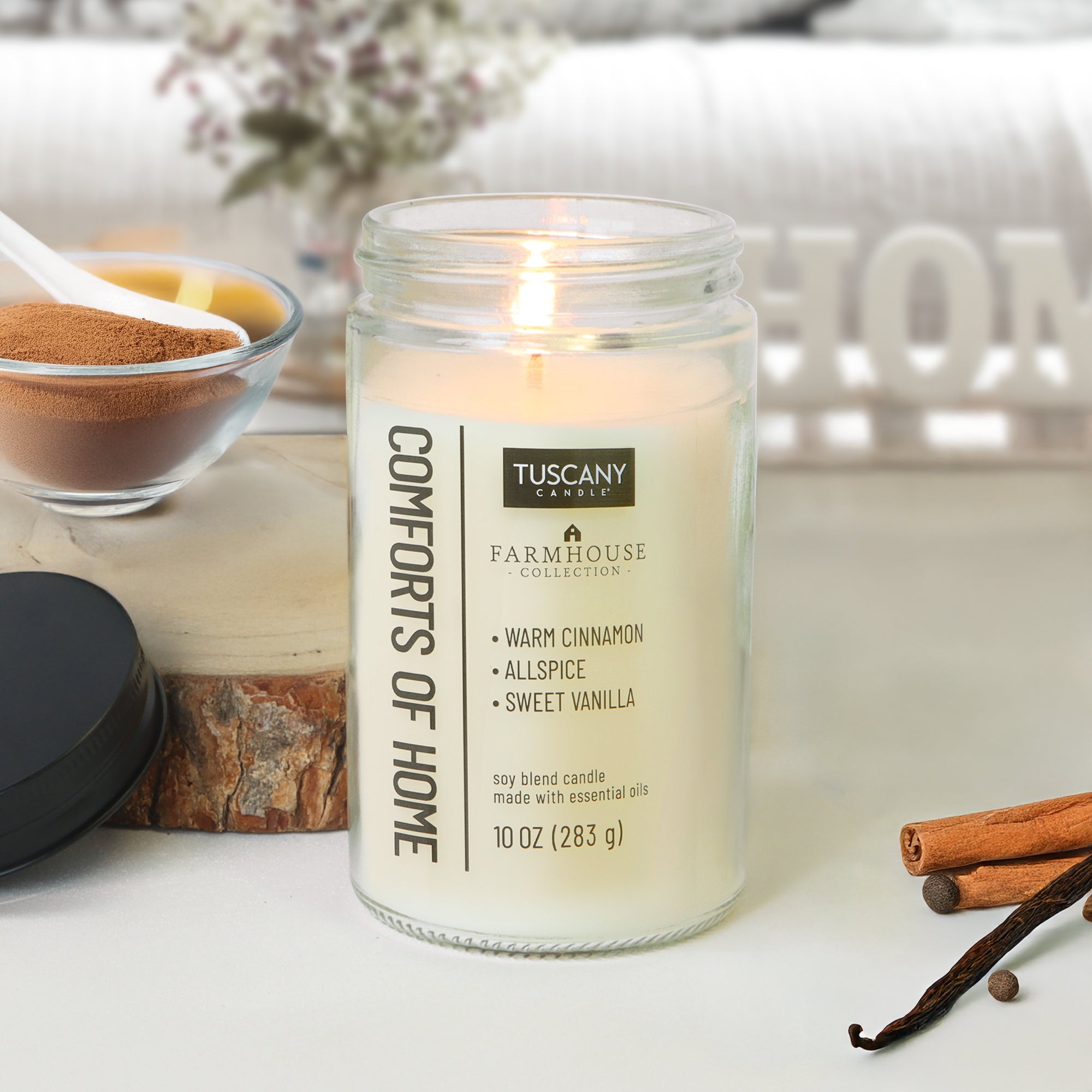 A Tuscany Candle® EVD Comforts of Home Scented Jar Candle (10 oz) from the Farmhouse Collection sits on a table with spices, a cinnamon stick, and peppercorns, filling the air with its cozy cinnamon vanilla aroma.