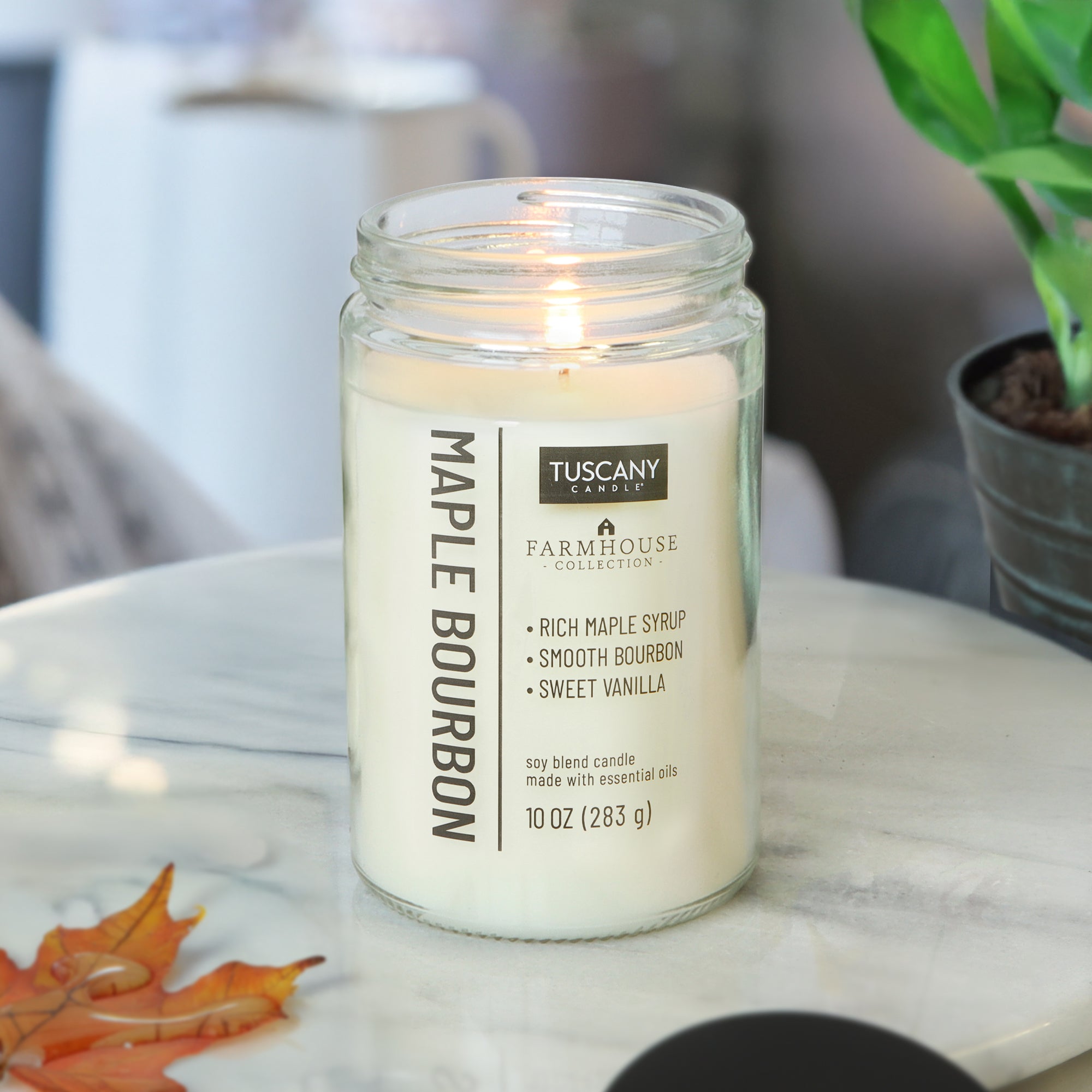A lit Maple Bourbon Scented Jar Candle (10 oz) from the Tuscany Candle® EVD Farmhouse Collection sits on a marble table near a potted plant and artificial autumn leaves, bringing rustic charm to the setting.