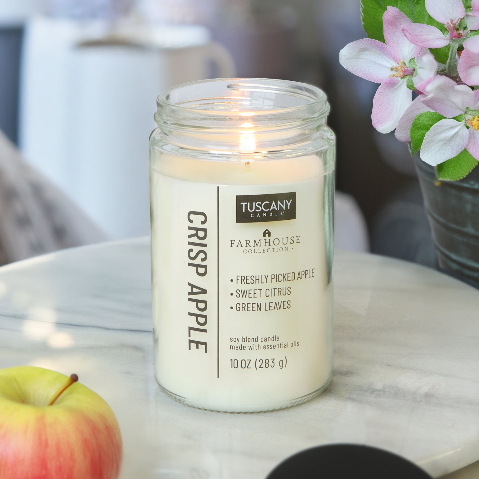 A lit Tuscany Candle® EVD Crisp Apple Scented Jar Candle (10 oz) from the Farmhouse Collection sits on a white surface beside a red-and-yellow apple and a flowering plant. The soy blend jar displays its scent notes and weight.