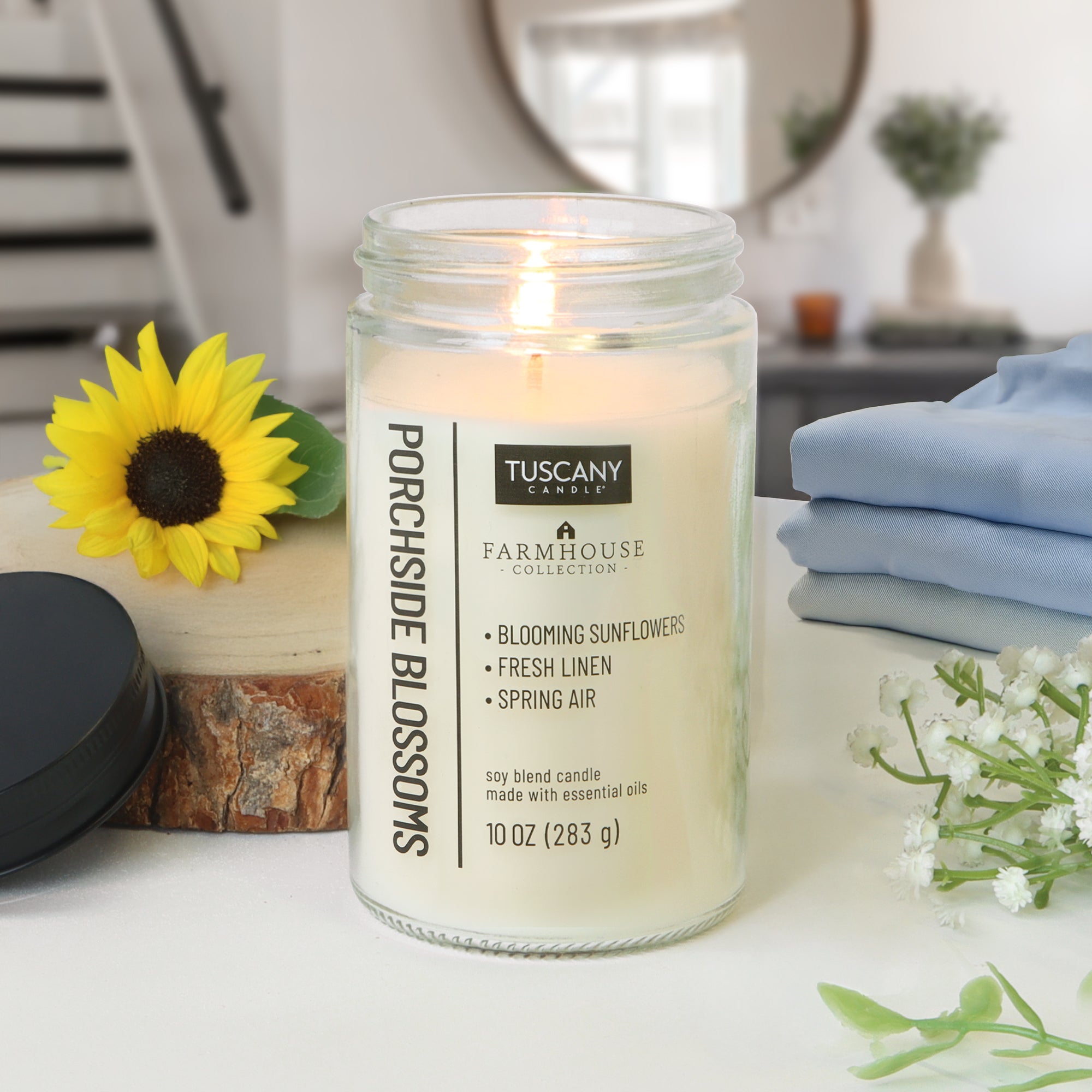 A lit Tuscany Candle® EVD Porchside Blossoms Scented Jar Candle (10 oz) from the Farmhouse Collection sits on a table near a sunflower, blue cloth, and white flowers, with a staircase and mirror in the background.