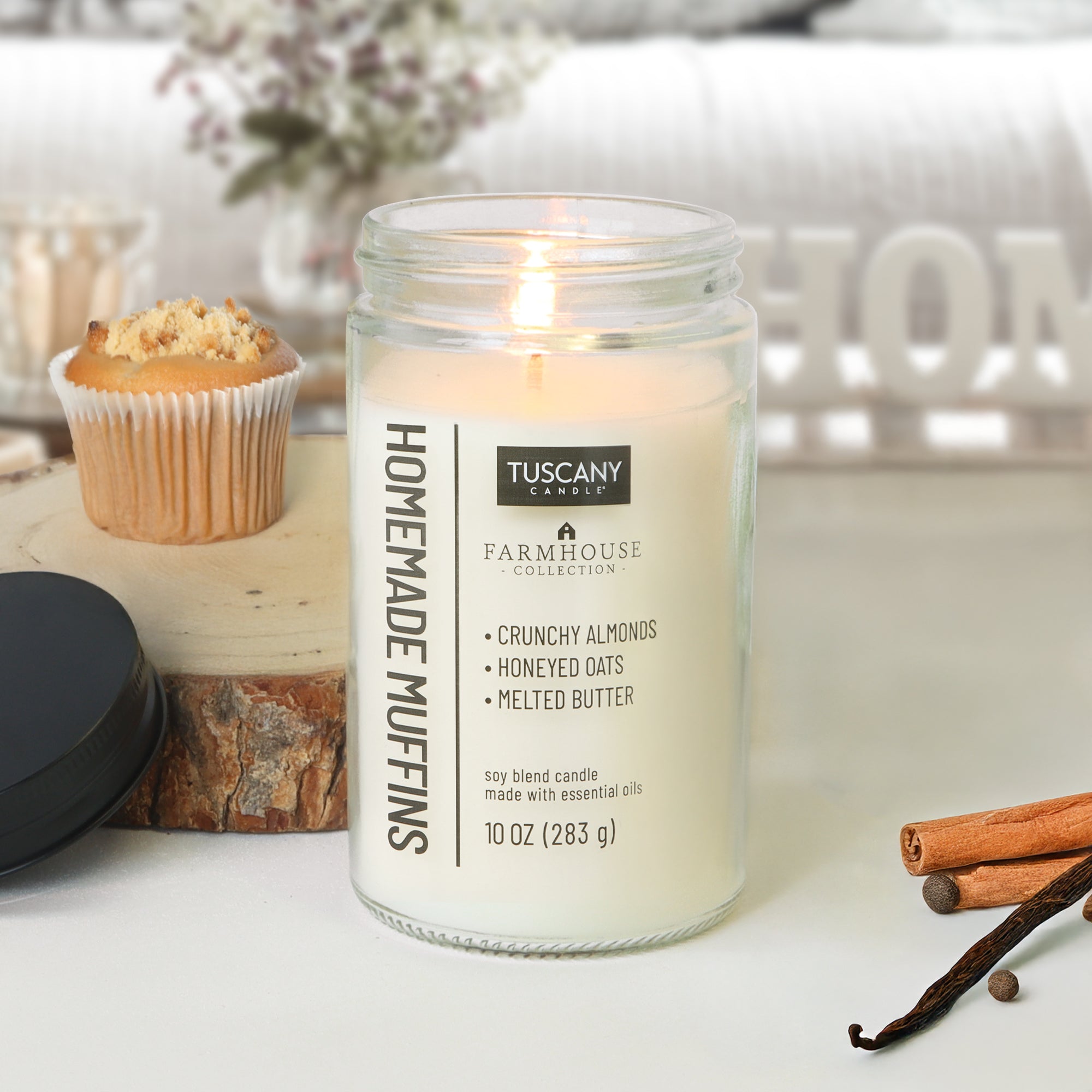 A Tuscany Candle® EVD Homemade Muffins Scented Jar Candle (10 oz) from the Farmhouse Collection sits on a table beside a muffin, cinnamon sticks, cloves, and its lid.