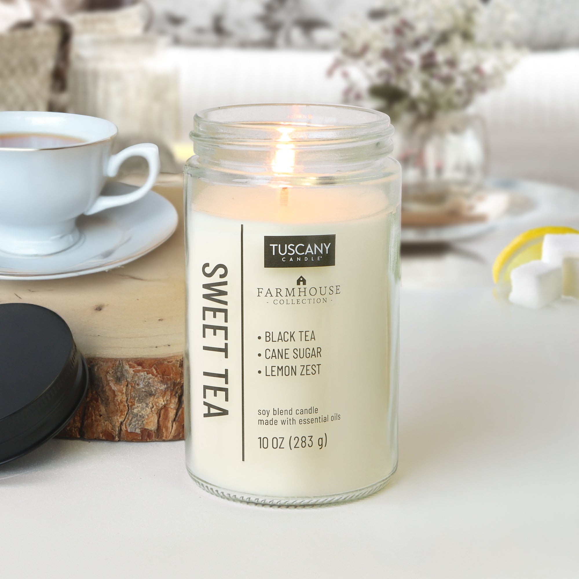 A lit Sweet Tea Scented Jar Candle (10 oz) from Tuscany Candle® EVD’s Farmhouse Collection fills the room with notes of black tea, cane sugar, and lemon zest, creating an inviting, cozy atmosphere near a teacup and wooden coaster.