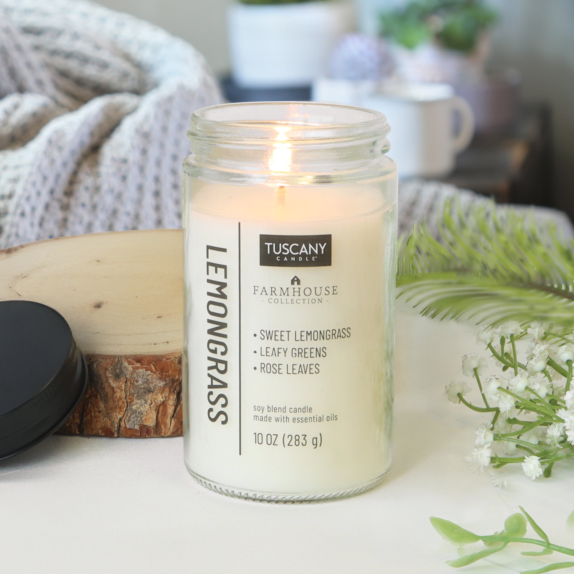 A 10 oz Lemongrass Scented Jar Candle from the Tuscany Candle® EVD Farmhouse Collection sits near a black lid amidst greenery and a white textured blanket, creating a cozy farmhouse vibe.