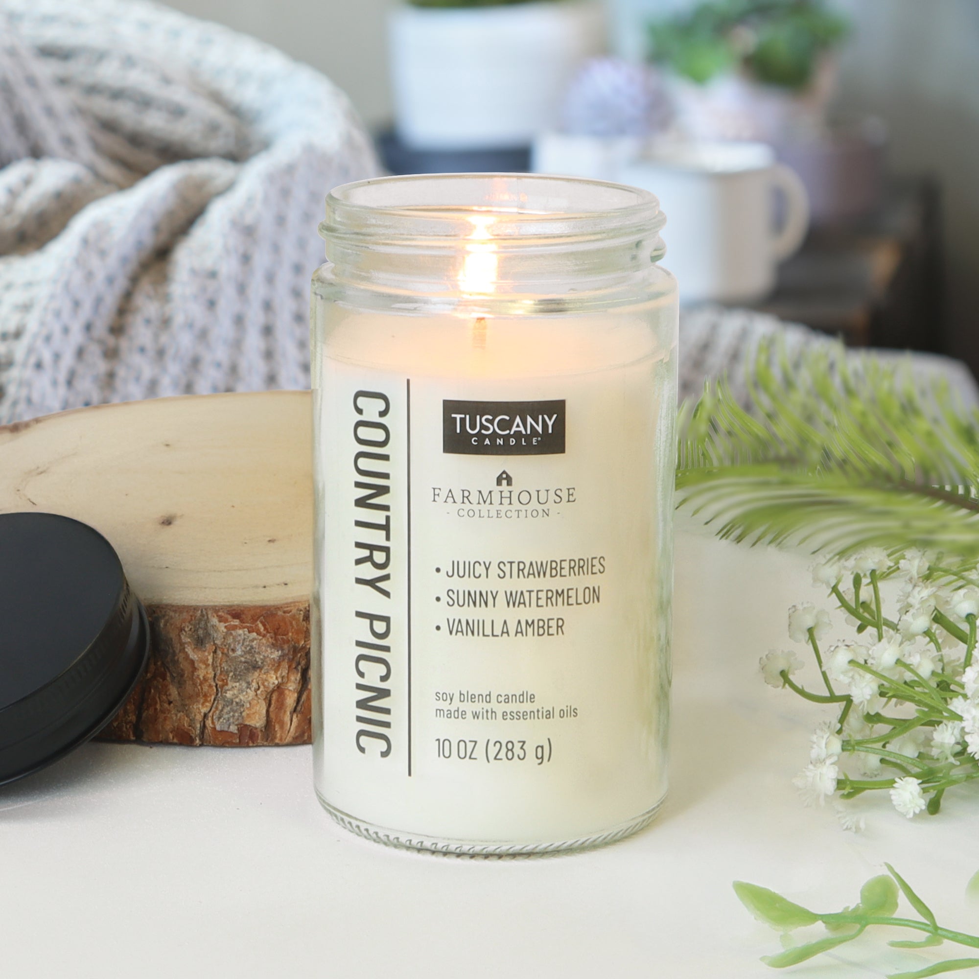 A Country Picnic Scented Jar Candle (10 oz) from Tuscany Candle® EVD sits with cozy décor, filling your space with strawberry and watermelon scents—the perfect farmhouse touch.