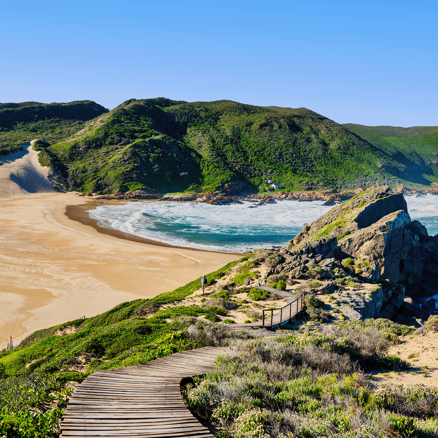 A wooden boardwalk leads to a sandy beach with green hills and cliffs beneath a blue sky, reflecting the fresh scent of Tuscany Candle® SEASONAL Seaside Orchid: Sea Breeze & Coastal Blooms Wax Melt—perfect for your coastal-inspired home.