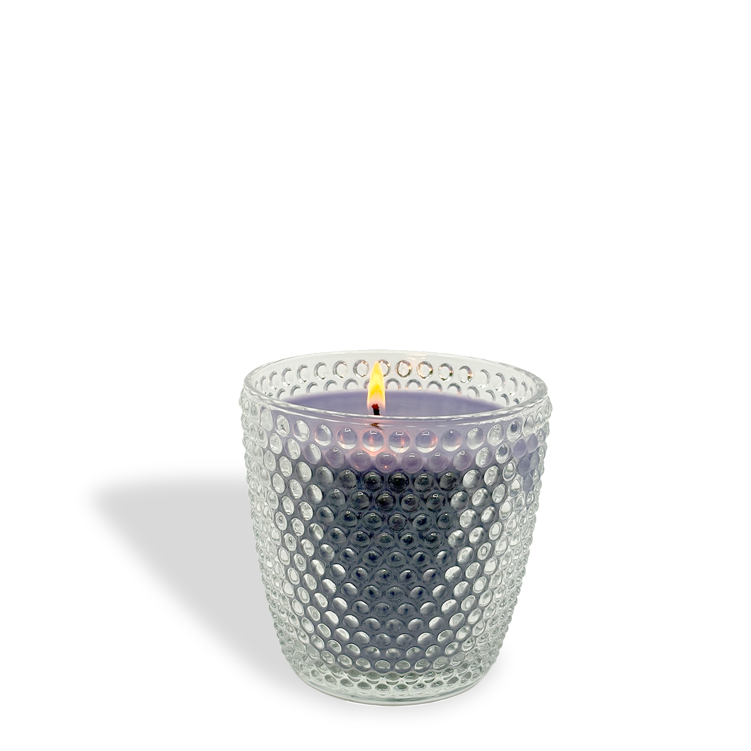 A lit Tuscany Candle® Lavender Lollipops: Grape & Jasmine Scented Easter Candle (8.5 oz) in a textured clear glass holder, grayish wax and visible flame, casts a soft shadow to the left.