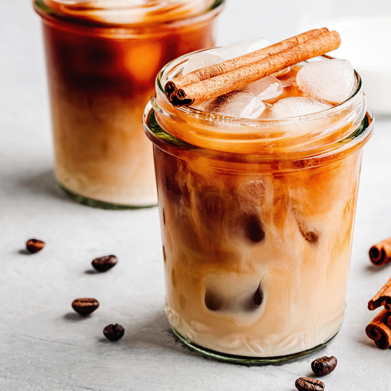 A glass of iced coffee with milk and ice, topped with cinnamon, sits beside coffee beans—mirroring the cozy aroma of Tuscany Candle® SEASONAL’s Iced Chai: Cardamom & Honey Scented Spring Candle (14 oz), made from a soy wax blend.