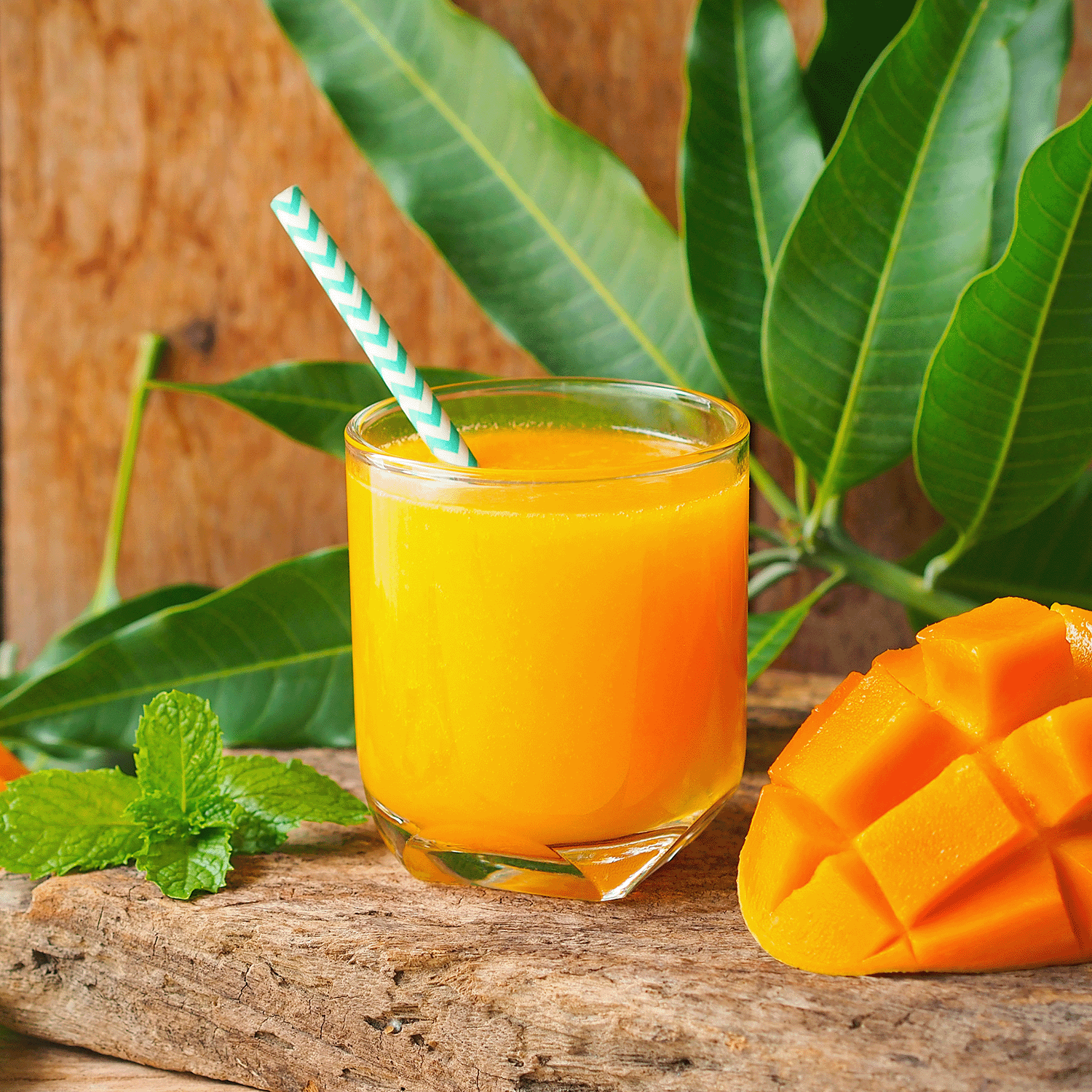 A glass of mango juice with a striped straw sits on wood beside sliced mango, mint, and the Tuscany Candle® SEASONAL Mango Blast: Pineapple & Peach Scented Spring Candle (14 oz), with green mango leaves in the background.