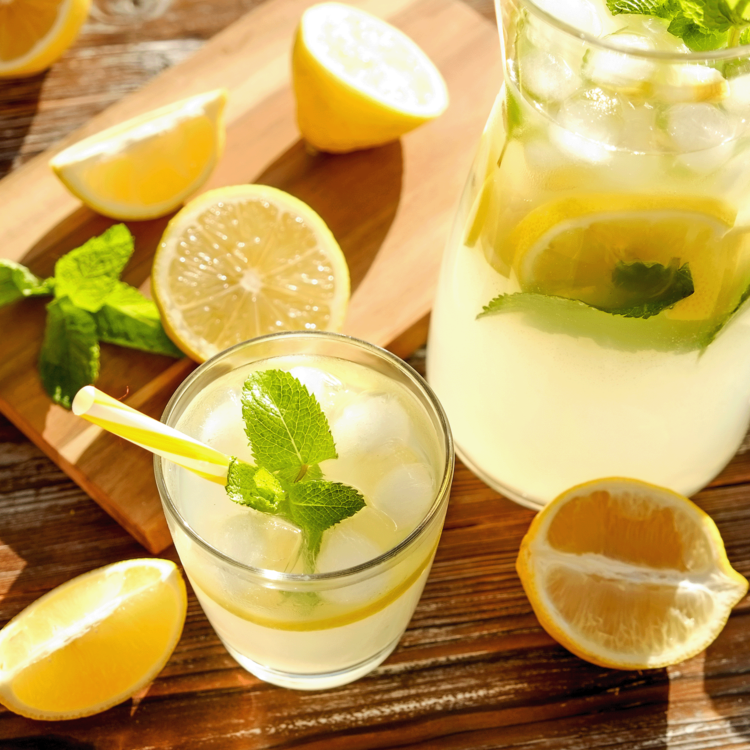 A glass and pitcher of lemonade with ice, lemon slices, and mint sit on a sunny wooden surface, capturing the zesty aroma of Tuscany Candle® SEASONAL Lemon Limeade: Sparkling Seltzer Scented Summer Candle (13 oz).