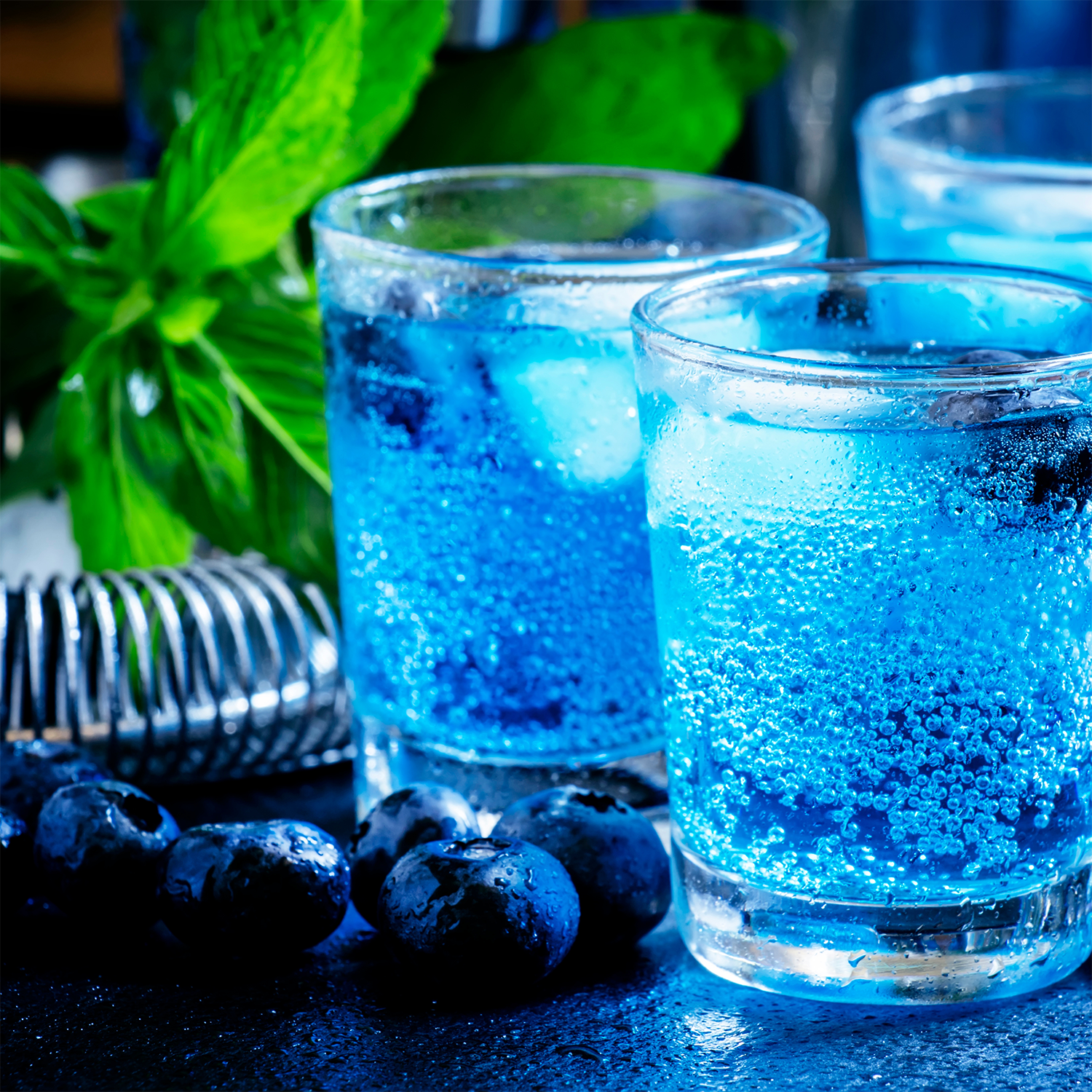 Two glasses of blue sparkling drink with ice, blueberries, and mint on a dark surface inspire the Tuscany Candle® SEASONAL Wildberry Fizz: Sparkling Seltzer Scented Summer Candle (13 oz)—the perfect summery home fragrance.