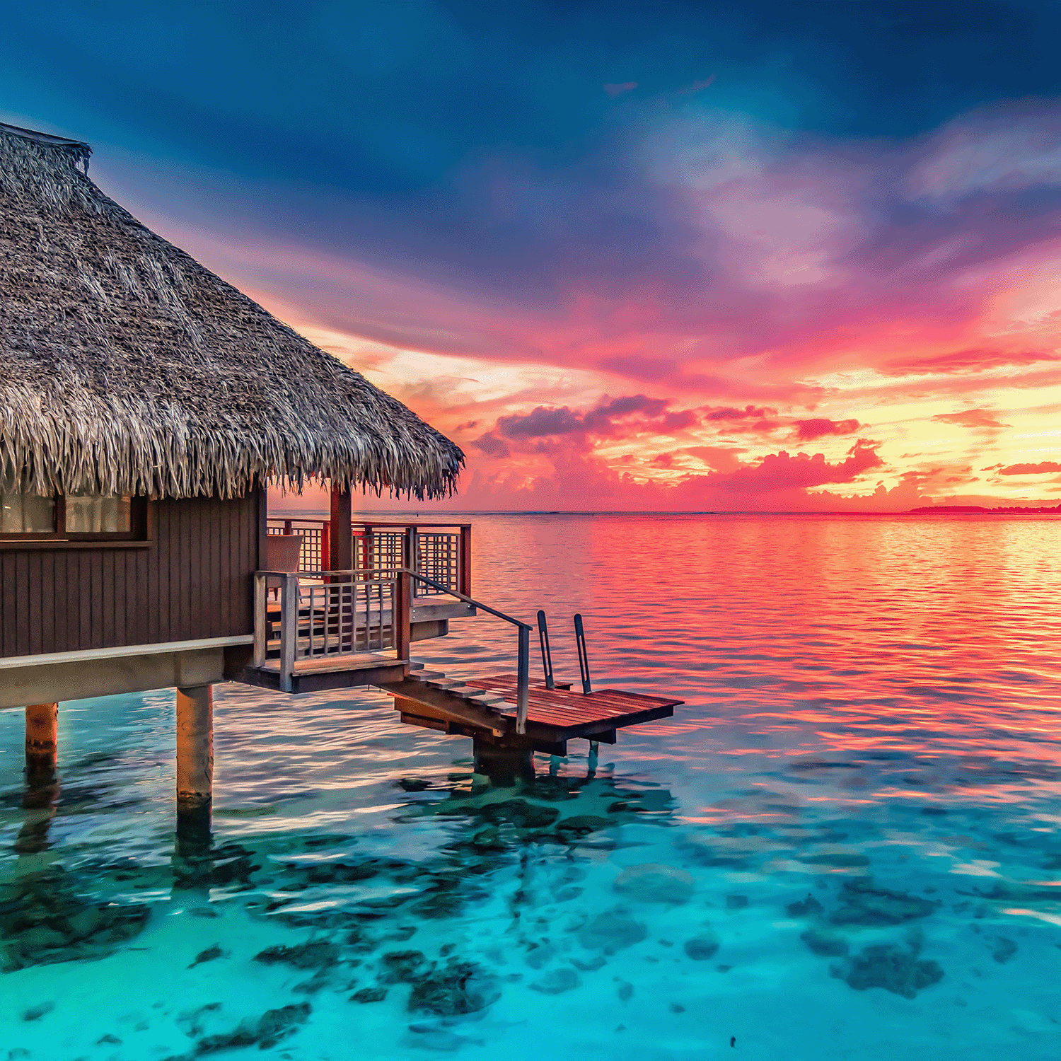A thatched-roof overwater bungalow faces a vibrant sunset as the Beachside Mangolada: Papaya & Mango Scented Spring Candle by Tuscany Candle® SEASONAL fills the air with tropical fragrance, blending with colors reflected on the sea.