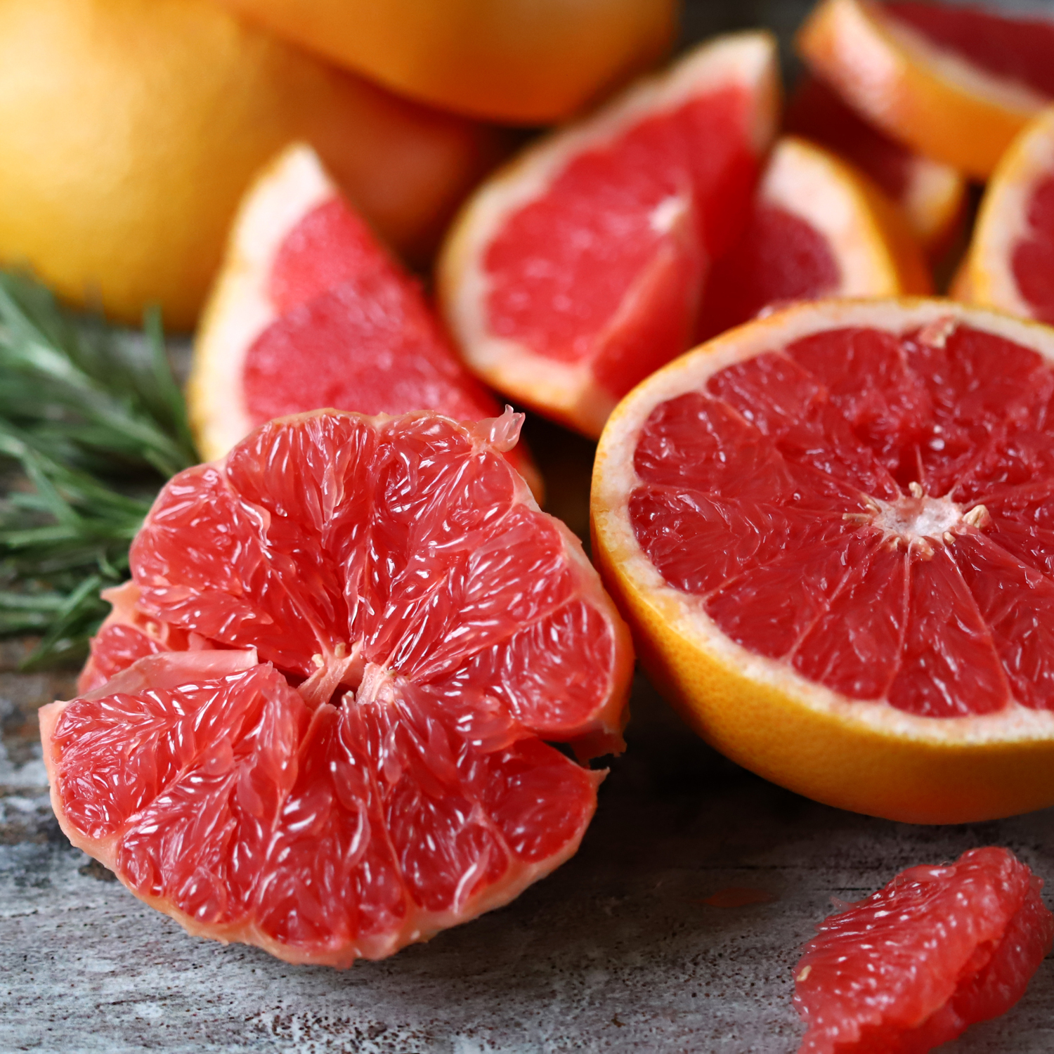Several ripe grapefruits, some halved and wedged, showcase their red flesh on rustic wood beside the Tuscany Candle® SEASONAL Amazing Mother: Grapefruit & Neroli Scented Spring Candle (11.5 oz), infusing the scene with color and fragrance.