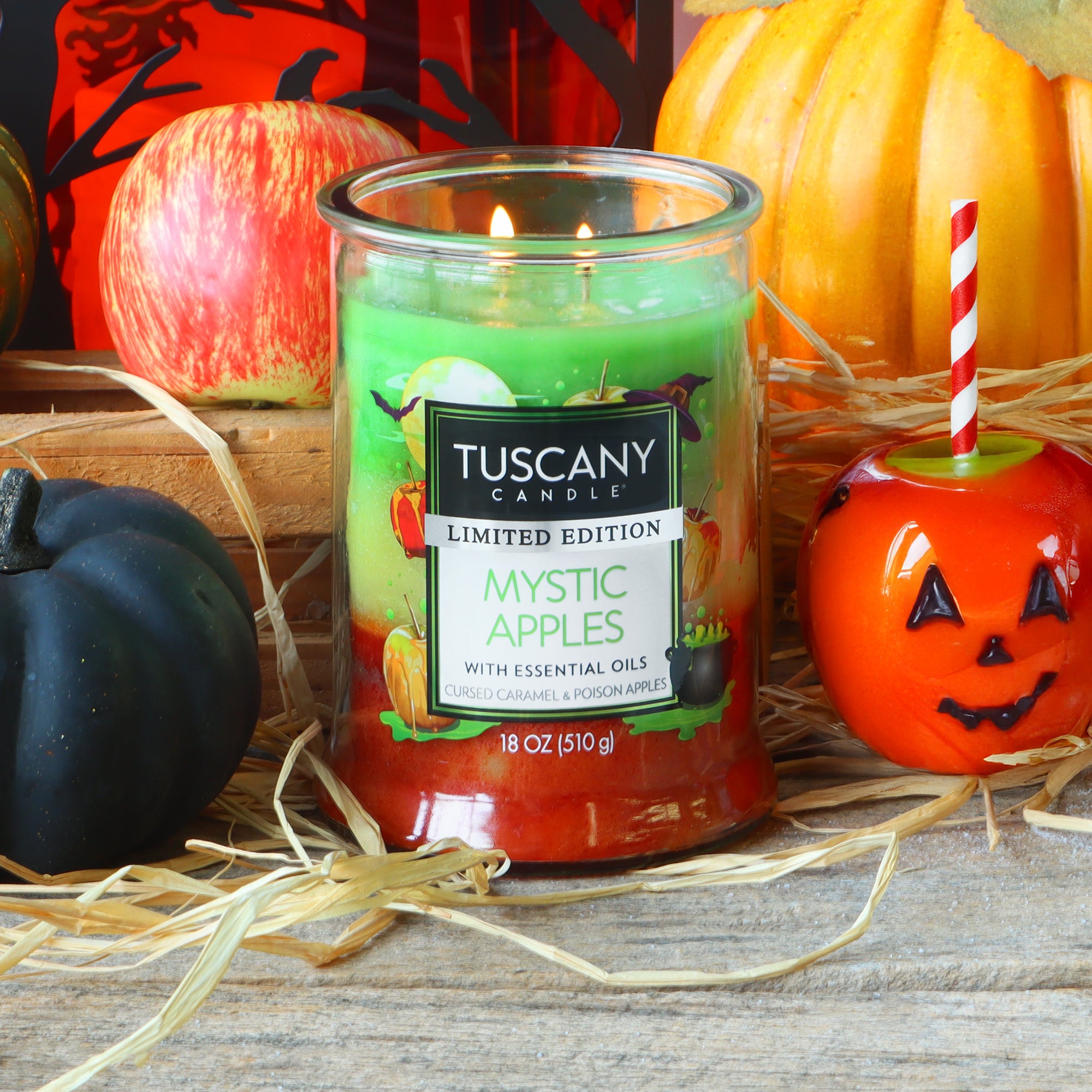 A Tuscany Candle® SEASONAL “Mystic Apples: Caramel & Poison Apples” scented Halloween candle (18 oz) burns beside black and orange pumpkins, a red candy apple, and straw on a wooden surface.