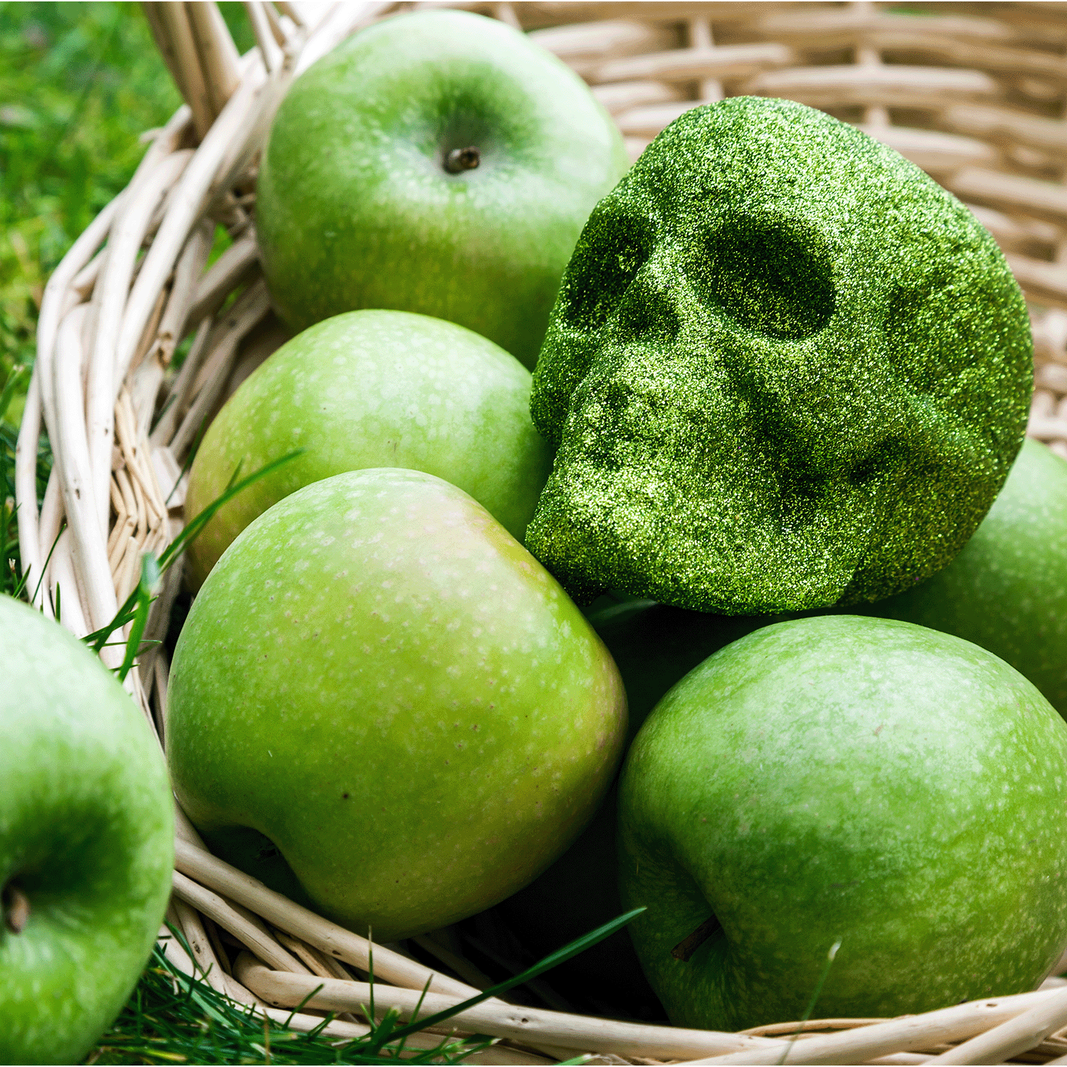 A wicker basket filled with Mystic Apples: Caramel & Poison Apples Scented Halloween Candle by Tuscany Candle® SEASONAL, surrounded by green apples and a green glittery skull decoration nestled among the fruit.