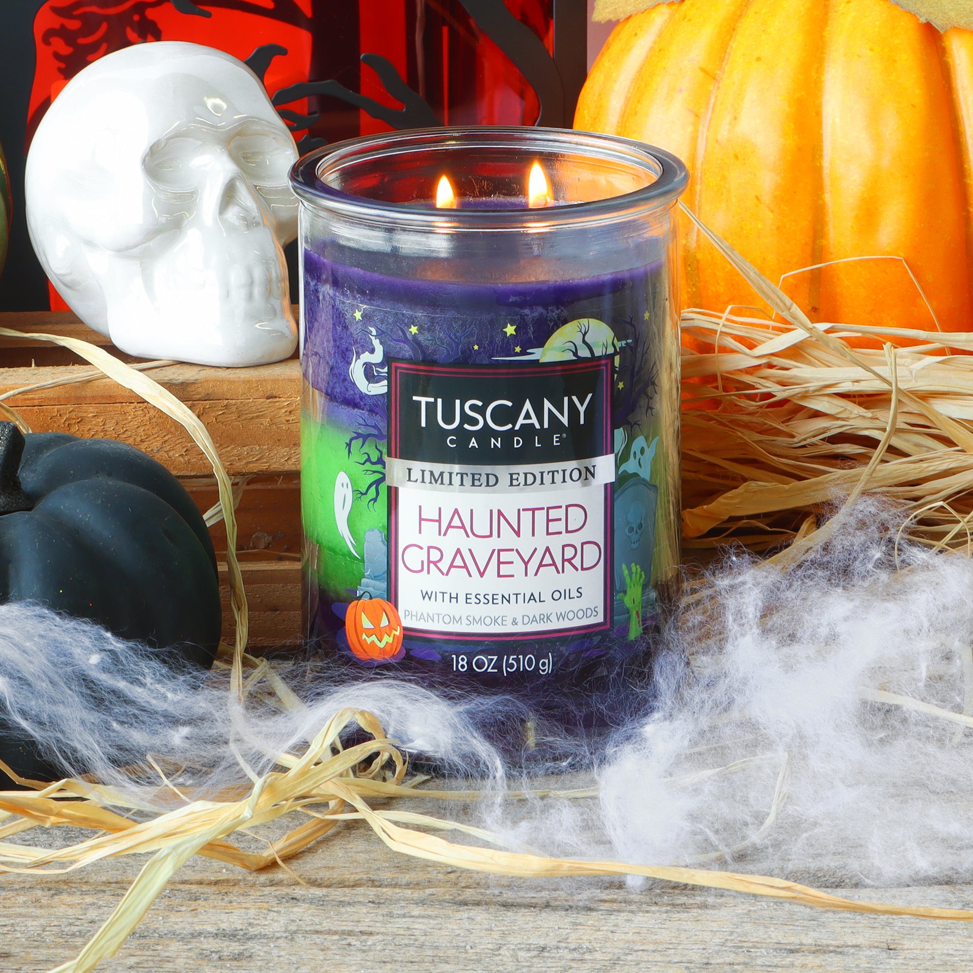 A Tuscany Candle® SEASONAL “Haunted Graveyard: Phantom Smoke & Dark Woods” scented Halloween candle (18 oz) burns on a wooden surface, surrounded by straw, spider web decorations, pumpkins, and other Halloween-themed decor.