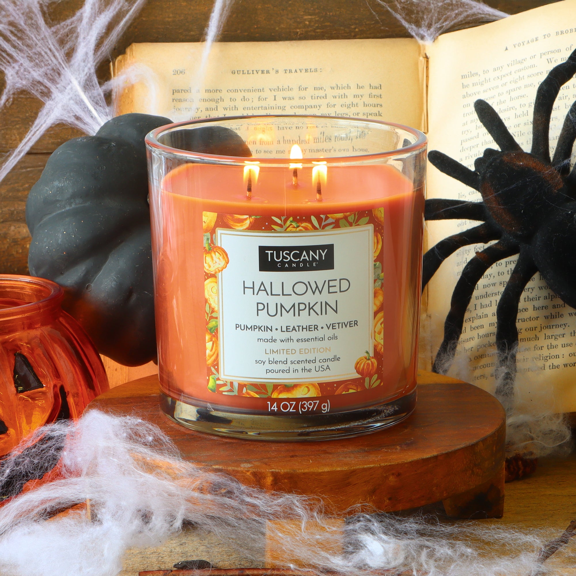 A lit Tuscany Candle® SEASONAL “Hallowed Pumpkin: Pumpkin & Leather Scented Halloween Candle” (14 oz) rests on a wooden surface, surrounded by artificial cobwebs, a black pumpkin, and a decorative spider.