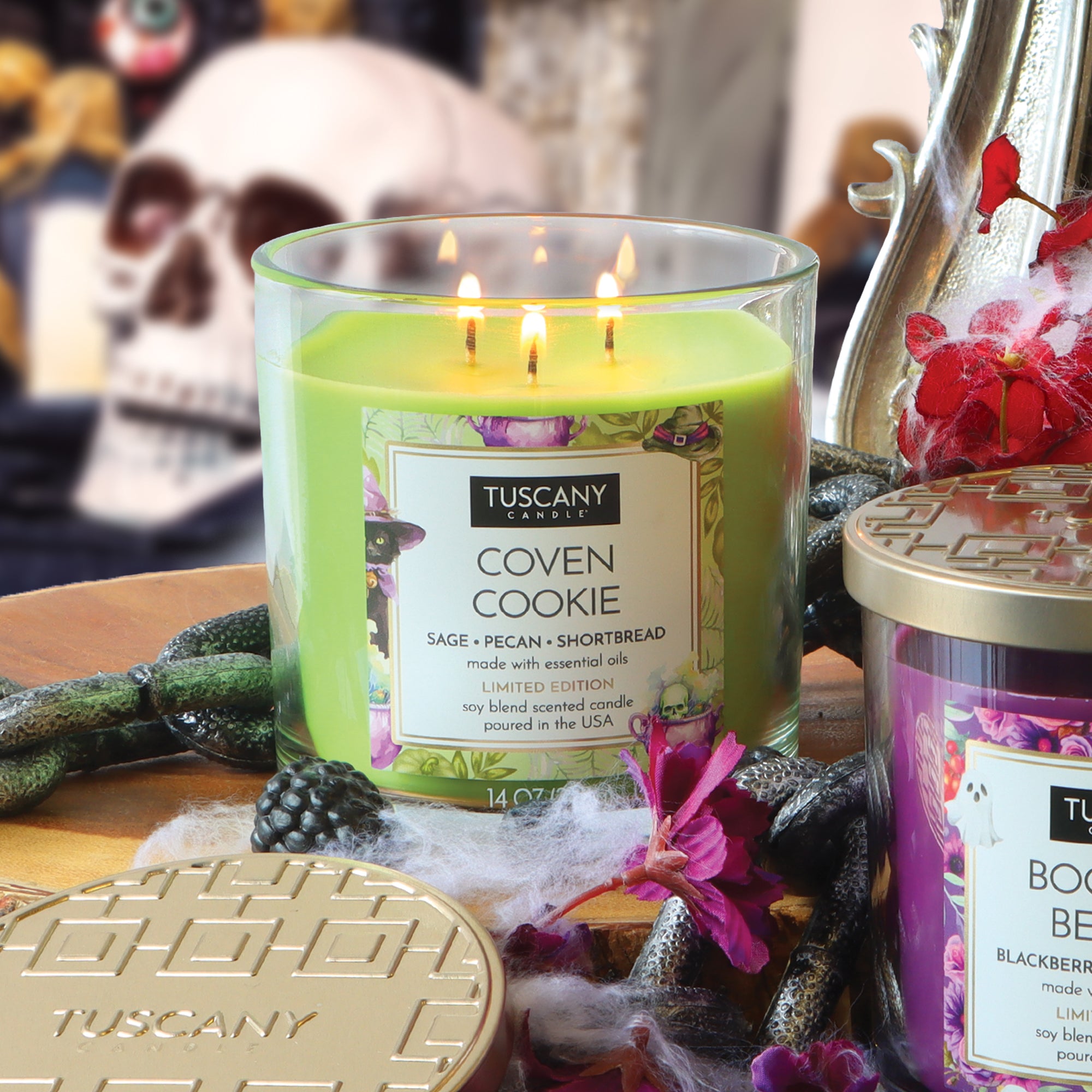 A green Tuscany Candle® SEASONAL “Coven Cookie: Pecan Shortbread Scented Halloween Candle” (14 oz) with three wicks burns brightly, surrounded by other candles, flowers, and festive Halloween décor in the background.