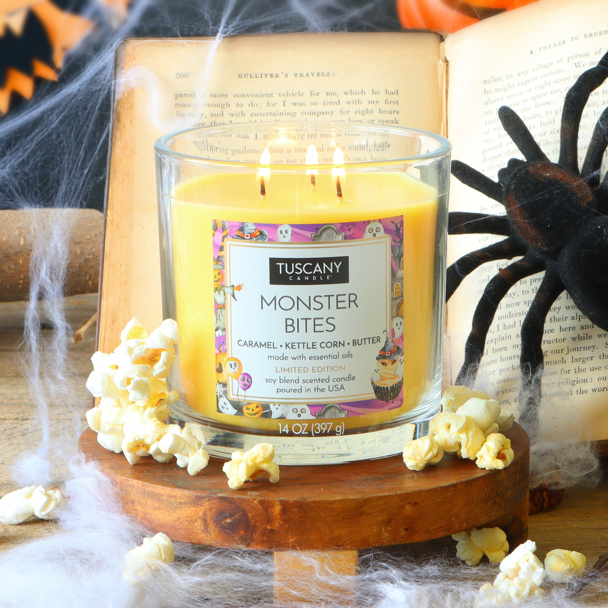 A lit Tuscany Candle® SEASONAL “Monster Bites: Caramel Kettlecorn Scented Halloween Candle” (14 oz) sits on a wooden surface, surrounded by popcorn, cobwebs, an open book, and a black spider decoration.