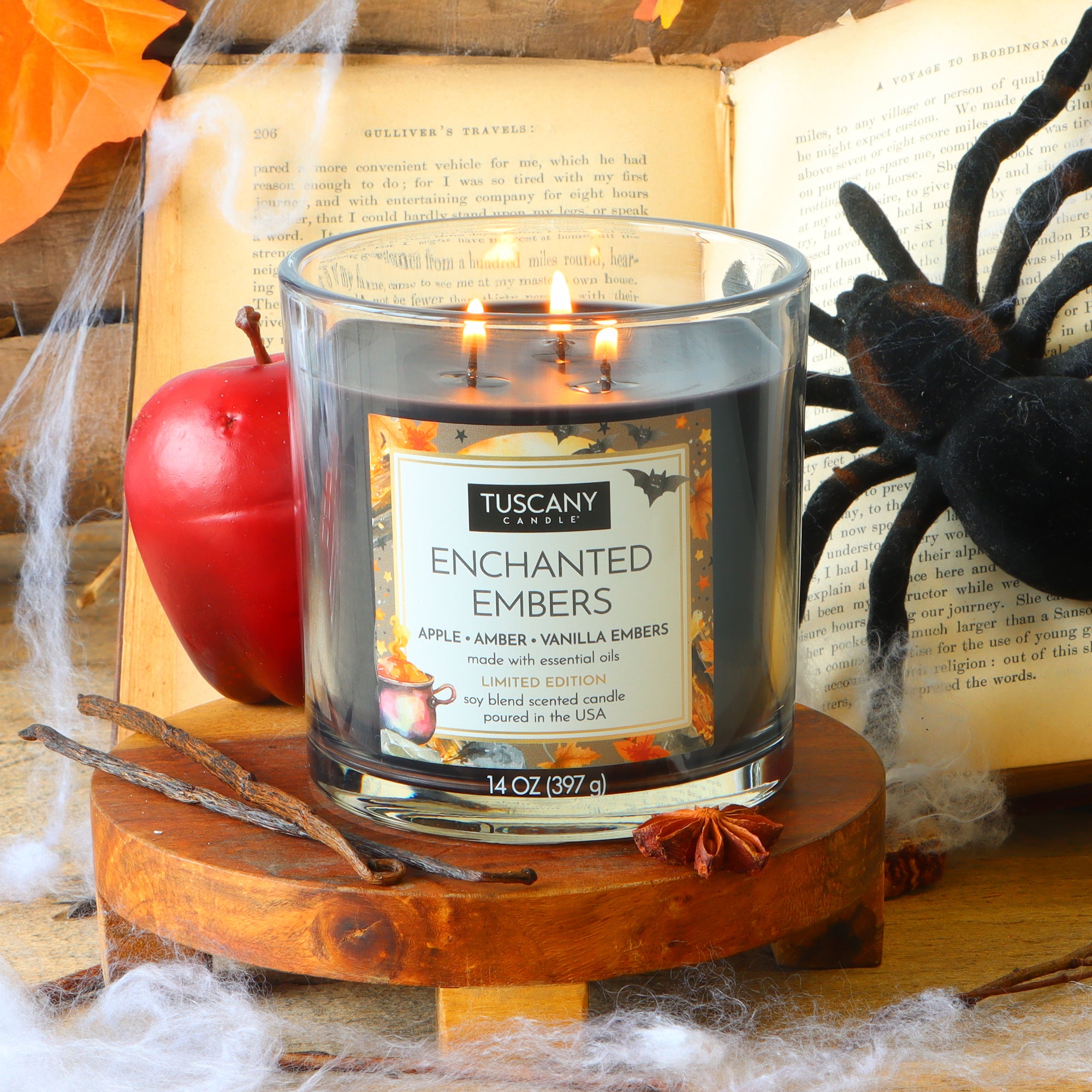 A Tuscany Candle® SEASONAL “Enchanted Embers: Apple & Amber Scented Halloween Candle” (14 oz) is displayed among fake cobwebs, a red apple, a plastic spider, cinnamon sticks, and an open book.