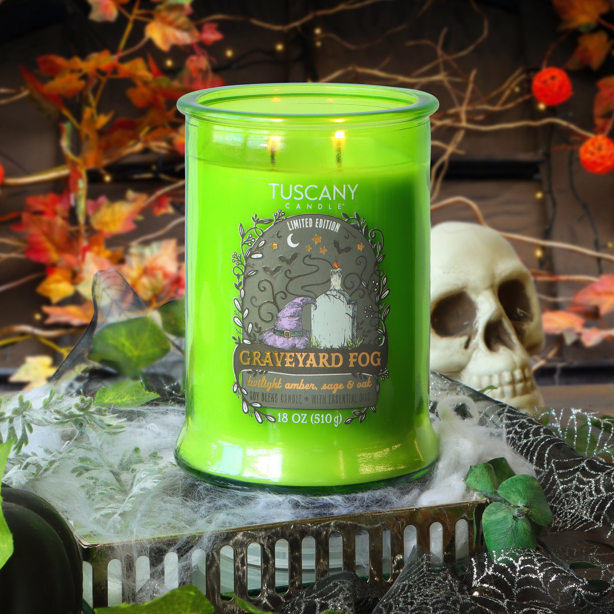 A green Tuscany Candle labeled “Graveyard Fog” burns on a table with faux cobwebs, leaves, a skull, and autumn decorations in the background.