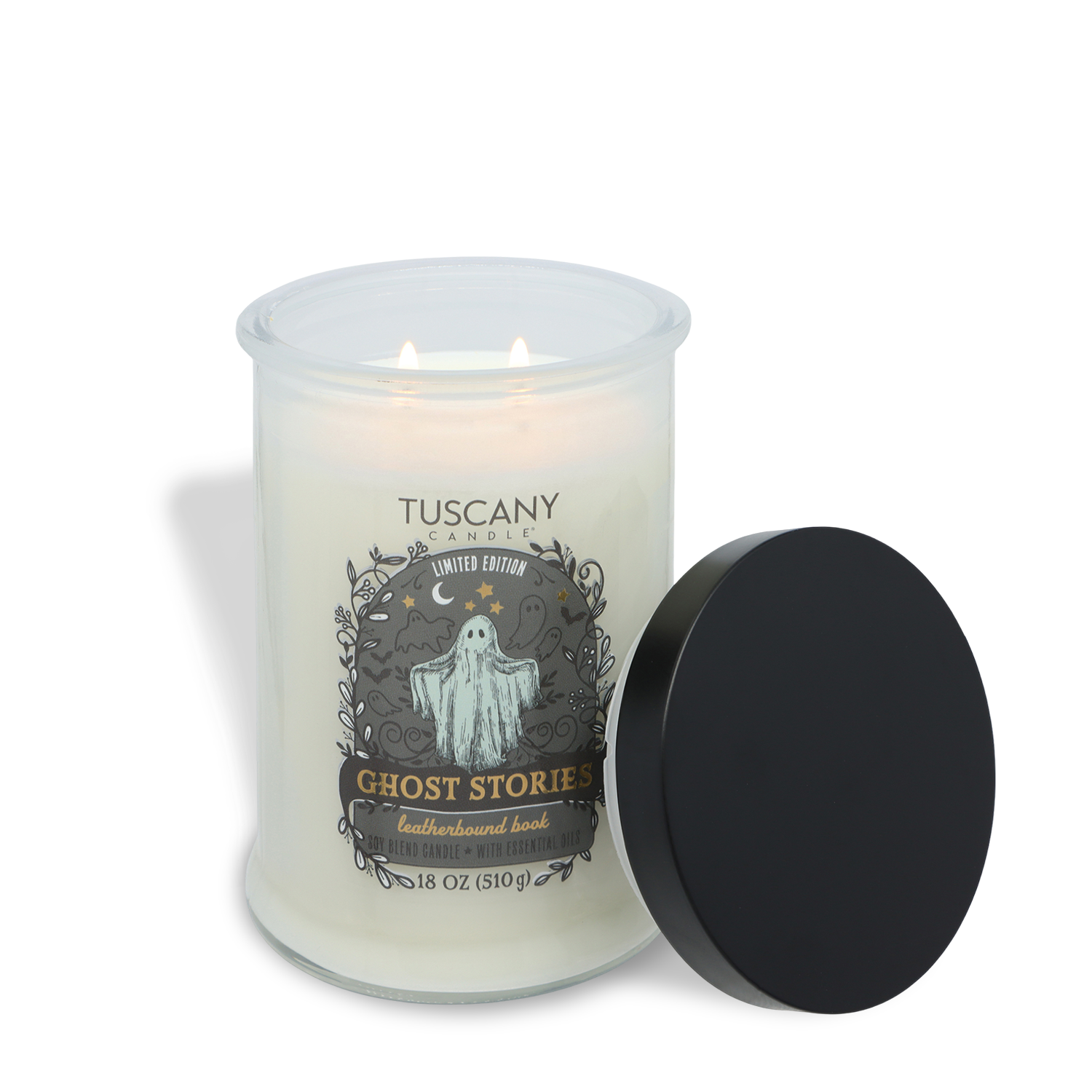 A white Tuscany Candle® SEASONAL candle labeled “Ghost Stories: Leatherbound Book Scented Halloween Candle” comes in a glass jar with a ghost illustration. The black lid is set beside the candle, and both wicks are lit.