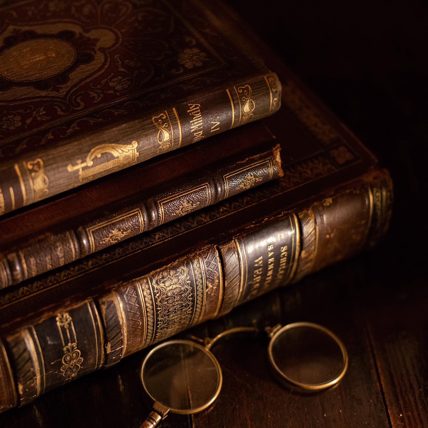 An 18 oz Ghost Stories: Leatherbound Book Scented Halloween Candle by Tuscany Candle® SEASONAL sits on a wooden surface next to a pair of round eyeglasses, its warm glow illuminating rich gold details reminiscent of antique book covers.
