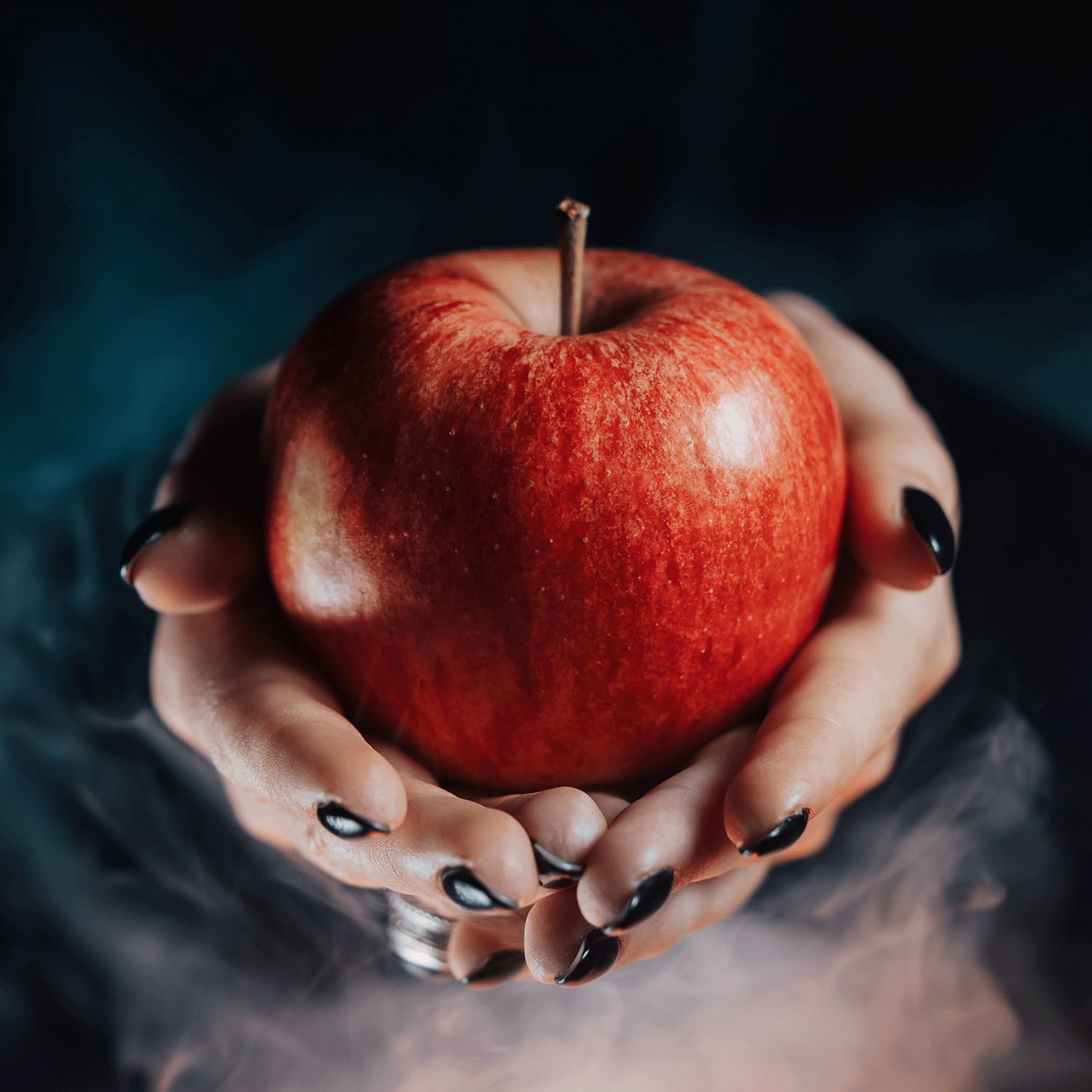 A pair of hands with black nail polish holds the Cursed Apple: Nutty Caramel Apple Scented Halloween Candle by Tuscany Candle® SEASONAL (12.5 oz), surrounded by wisps of smoke against a dark background.