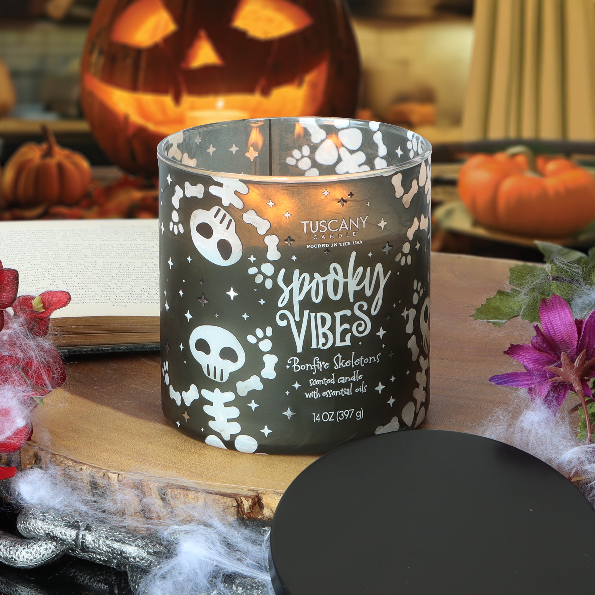 A lit Tuscany Candle® SEASONAL “Spooky Vibes: Bonfire Skeletons” scented Halloween candle (14 oz) sits on a wooden surface, surrounded by pumpkins and fake cobwebs as festive Halloween decorations.