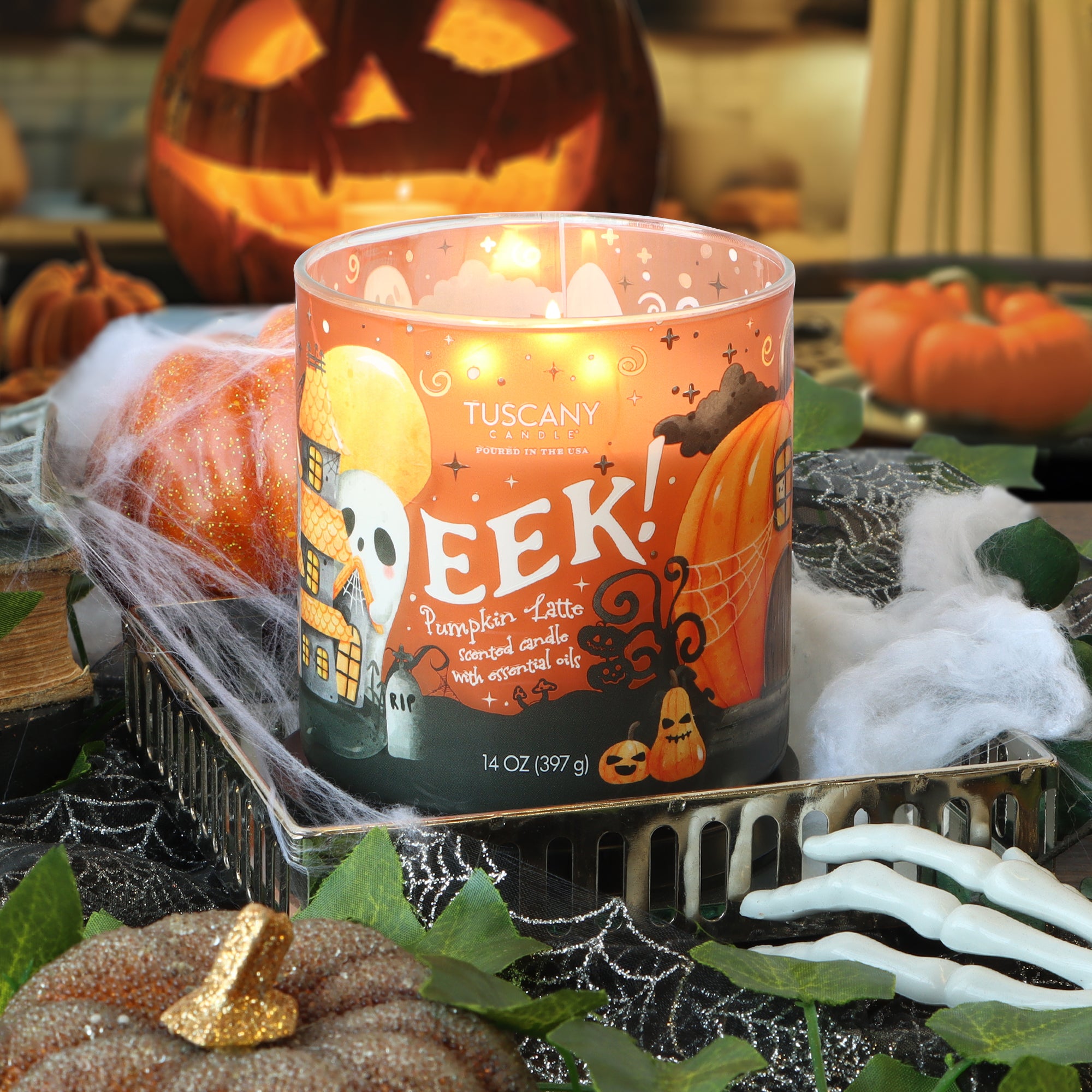 A Tuscany Candle® SEASONAL Eek: Pumpkin Latte Scented Halloween Candle (14 oz) burns on a tray surrounded by pumpkins, a skeleton hand, leaves, and a jack-o’-lantern in the background.