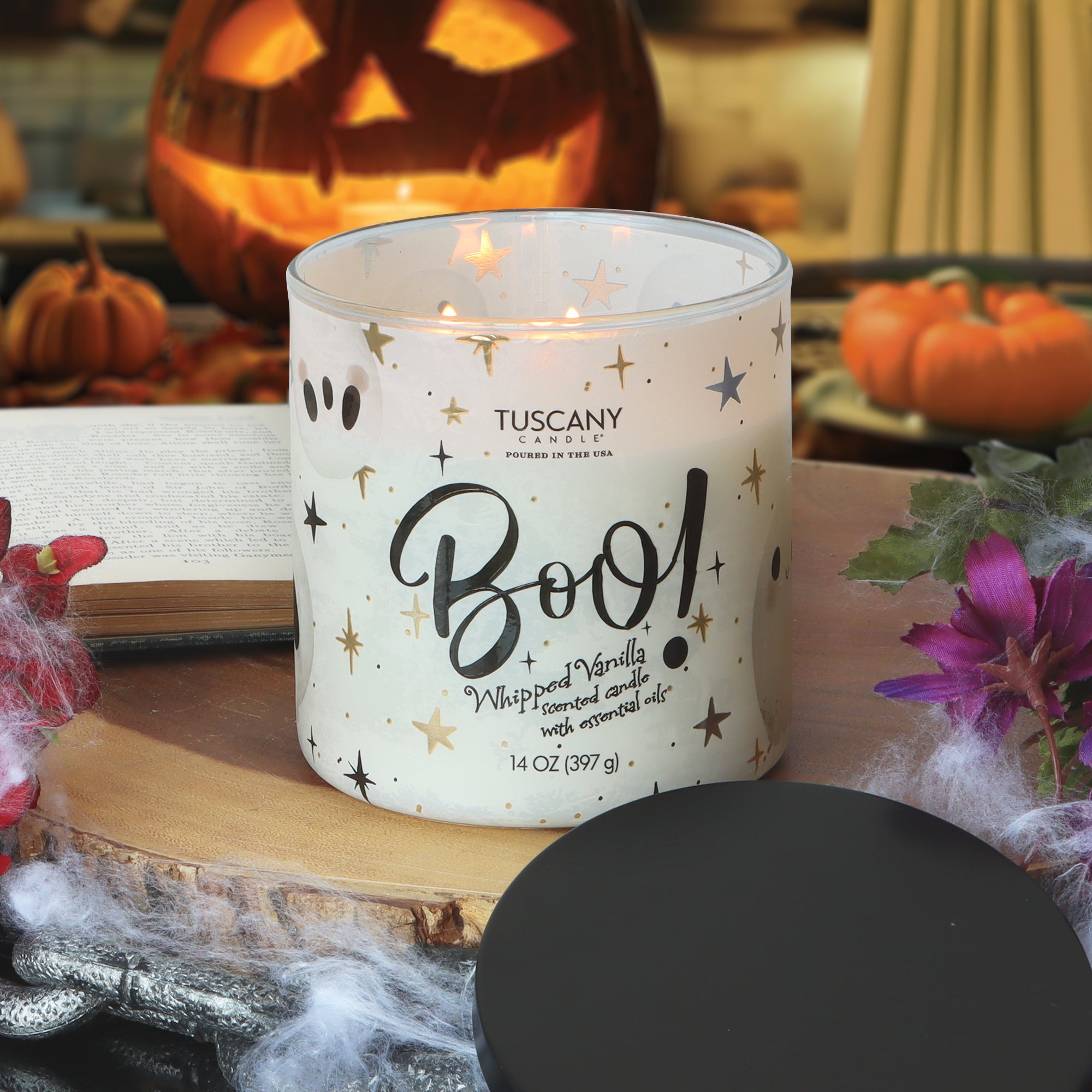 A Tuscany Candle® SEASONAL Boo: Whipped Vanilla Scented Halloween Candle (14 oz) gleams on a table surrounded by flowers, cobwebs, pumpkins, and a carved jack-o’-lantern in the background.