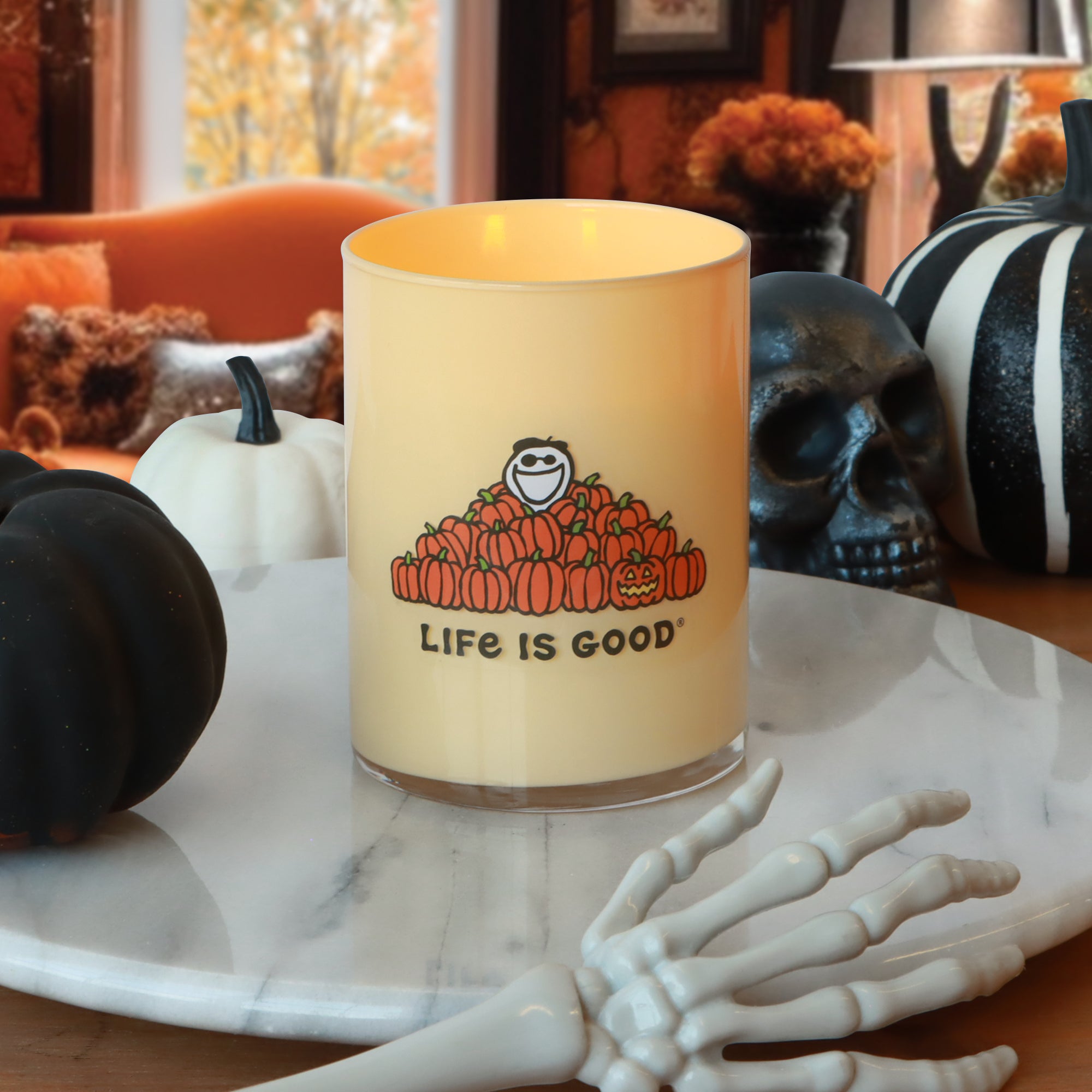A 13 oz Life Is Good: Vanilla Pumpkin Scented Halloween Candle by Tuscany Candle® SEASONAL, featuring “Life is Good” and pumpkin graphics, is displayed on a marble tray alongside black and white pumpkins, a skull, and a skeleton hand in a fall-themed room.