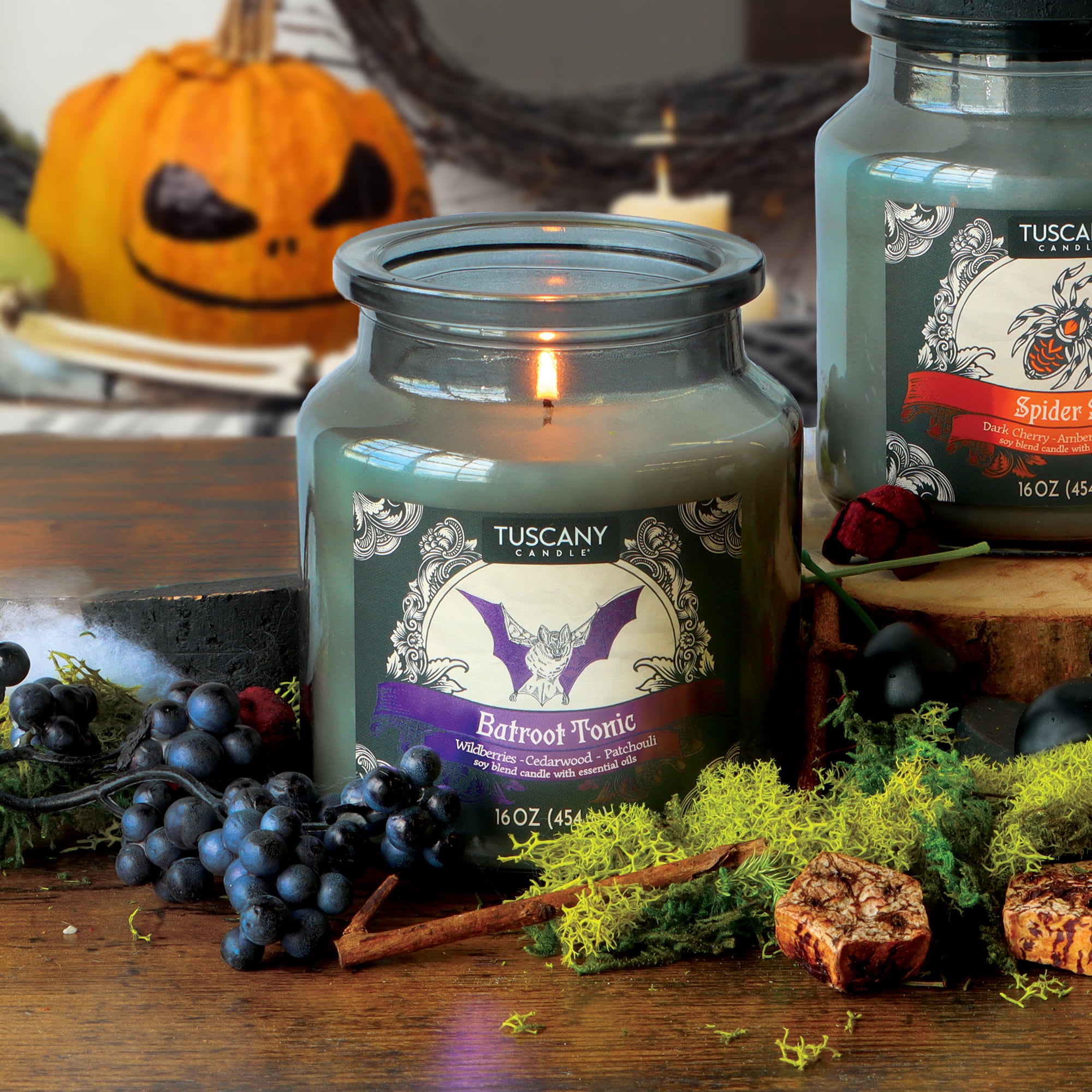 A Tuscany Candle® SEASONAL “Batroot Tonic: Wildberries & Cedarwood Scented Halloween Candle” (16 oz) rests on a wooden table accented with moss, berries, and a carved pumpkin in the background.