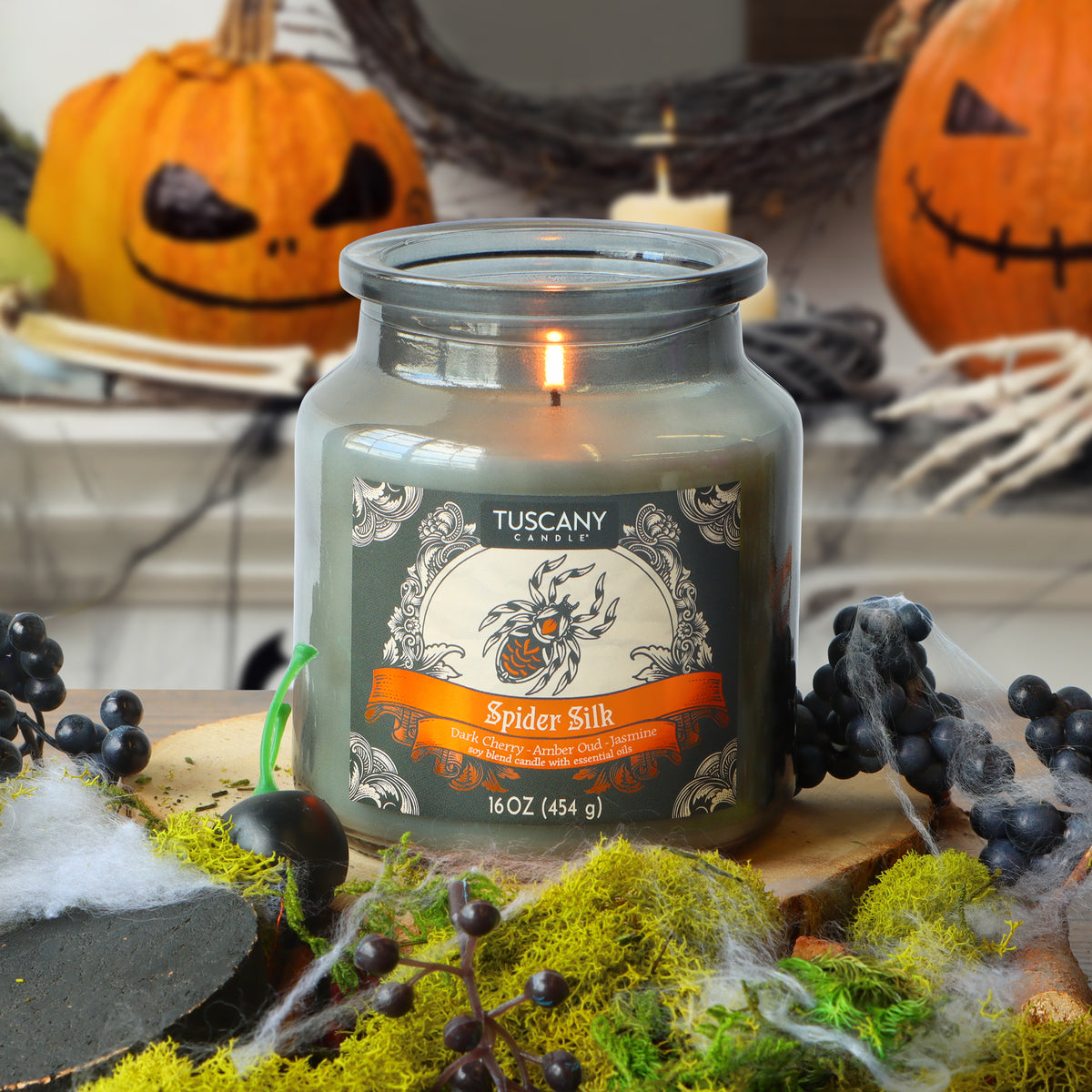 A Tuscany Candle® SEASONAL “Spider Silk: Dark Cherry & Amber Oud Scented Halloween Candle” (16 oz) burns on a table adorned with moss, faux spider webs, and black berries, while two carved pumpkins can be seen in the background.